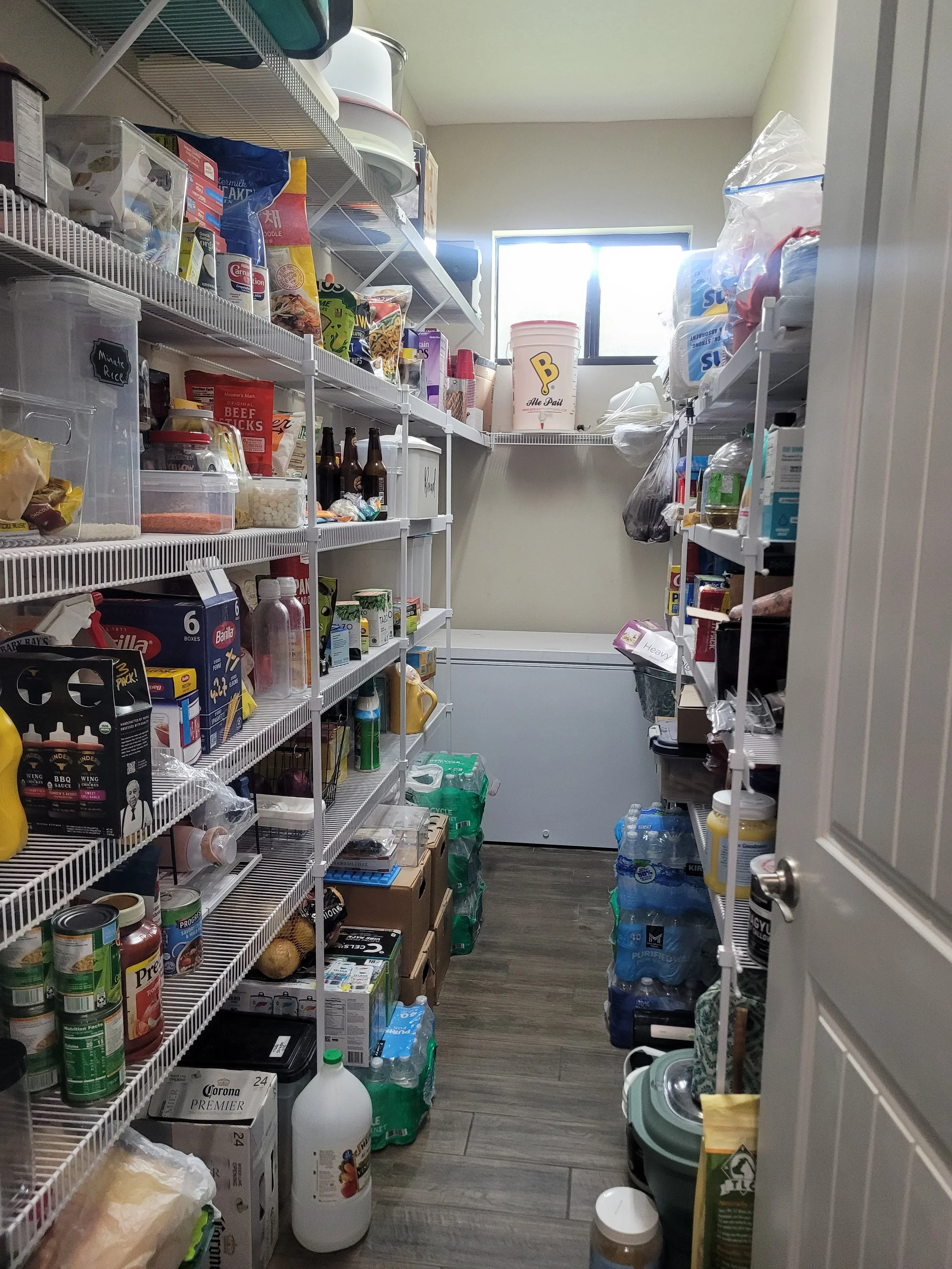 A small pantry with shelves filled with various food and drink items, including canned goods, bottles, and boxes, with a window at the back showing bright sunlight.