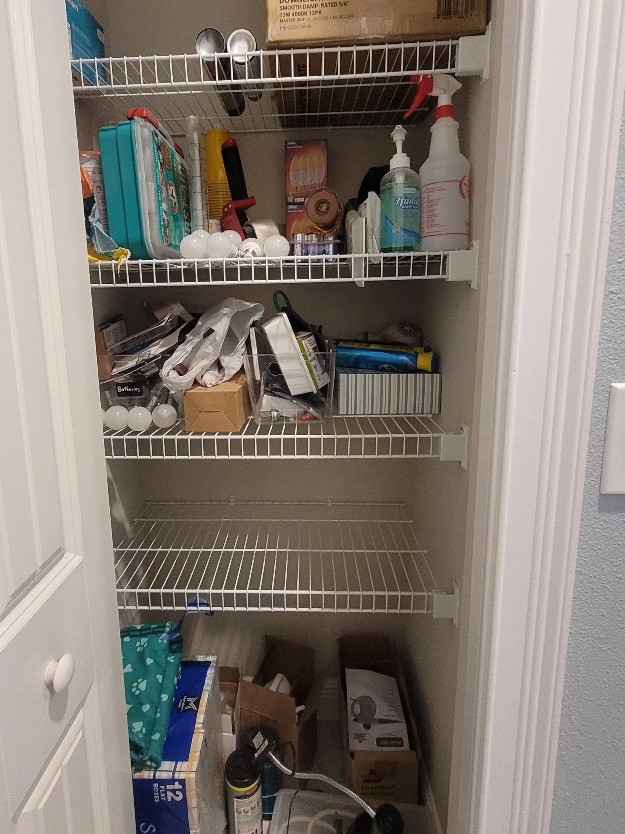 A cluttered closet with wire shelves filled with various household items including light bulbs, cleaning supplies, boxes, and miscellaneous objects.