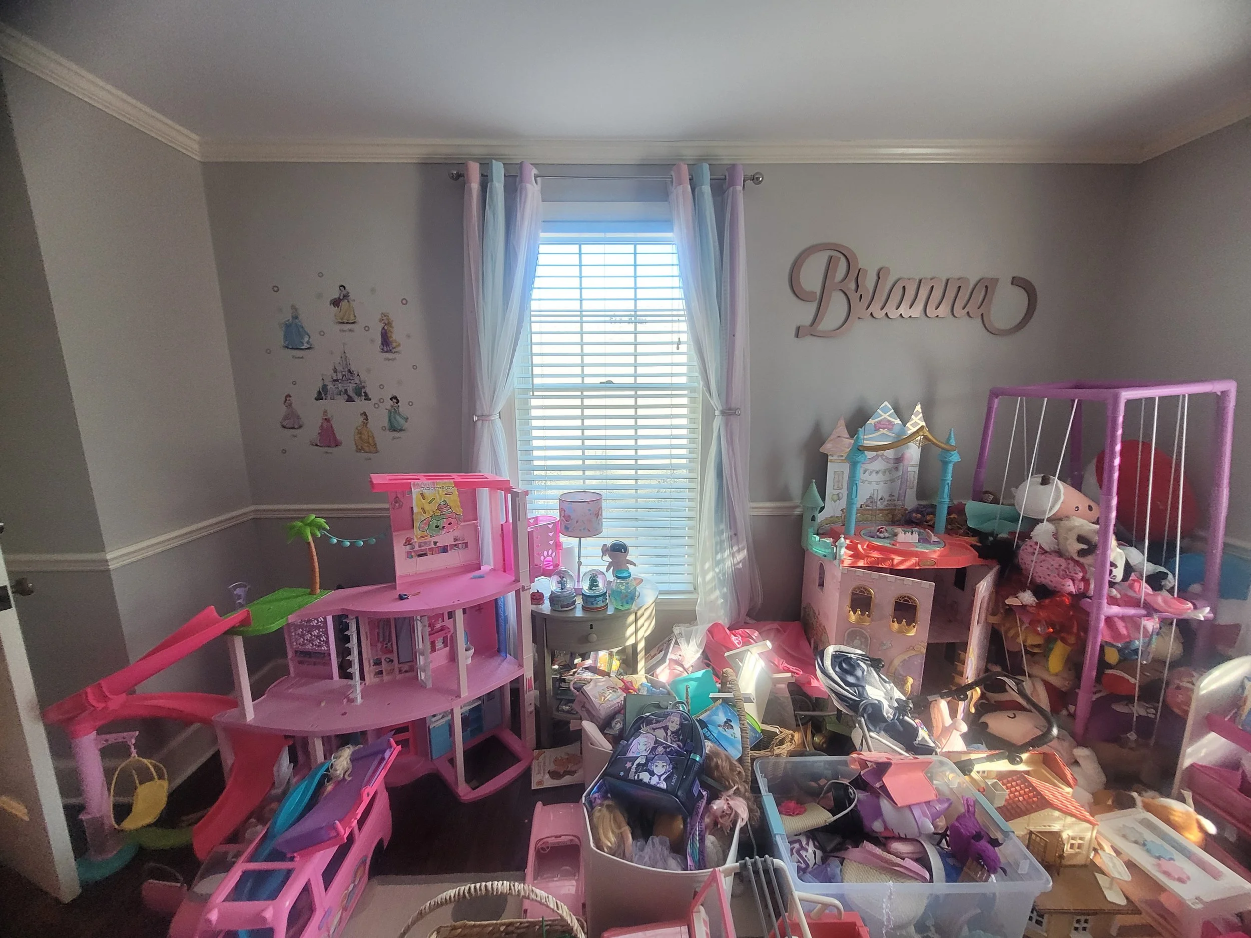 A cluttered children's room filled with various toys, dolls, and play furniture, with a window covered by white blinds and pastel-colored curtains, and VWanna written on the wall.