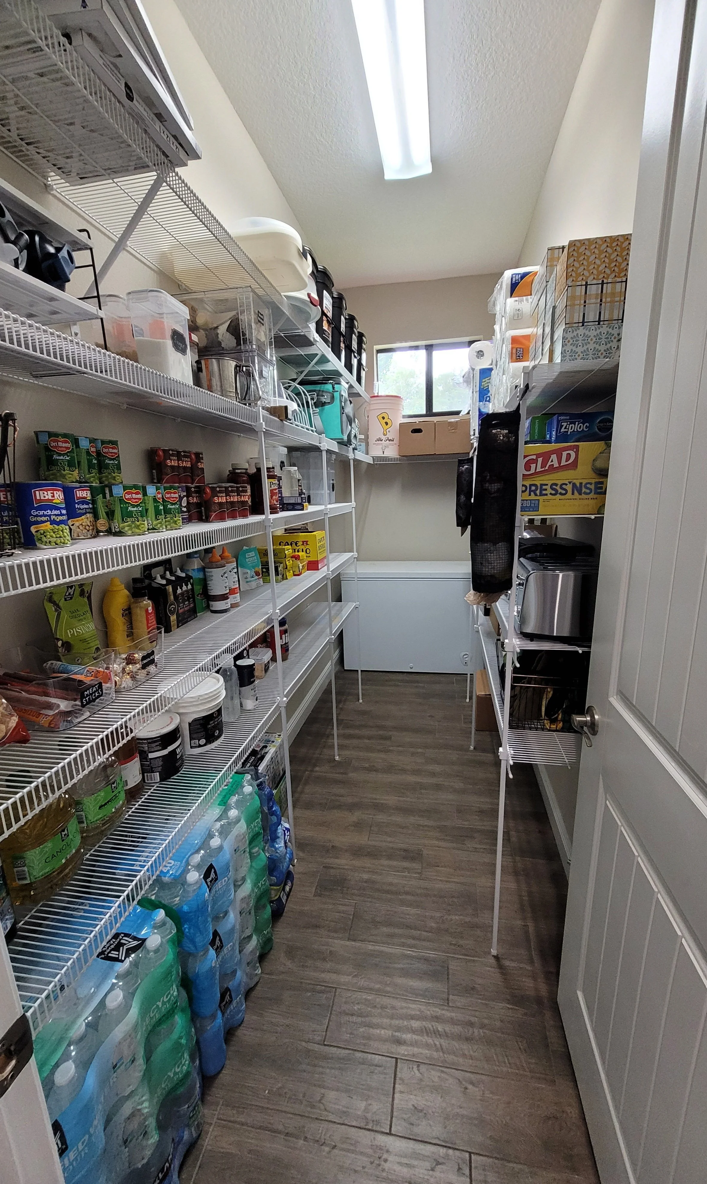 A walk-in pantry with wired shelves filled with canned goods, bottled water, cleaning supplies, and various packaged foods, with a small window at the back and a ceiling light.