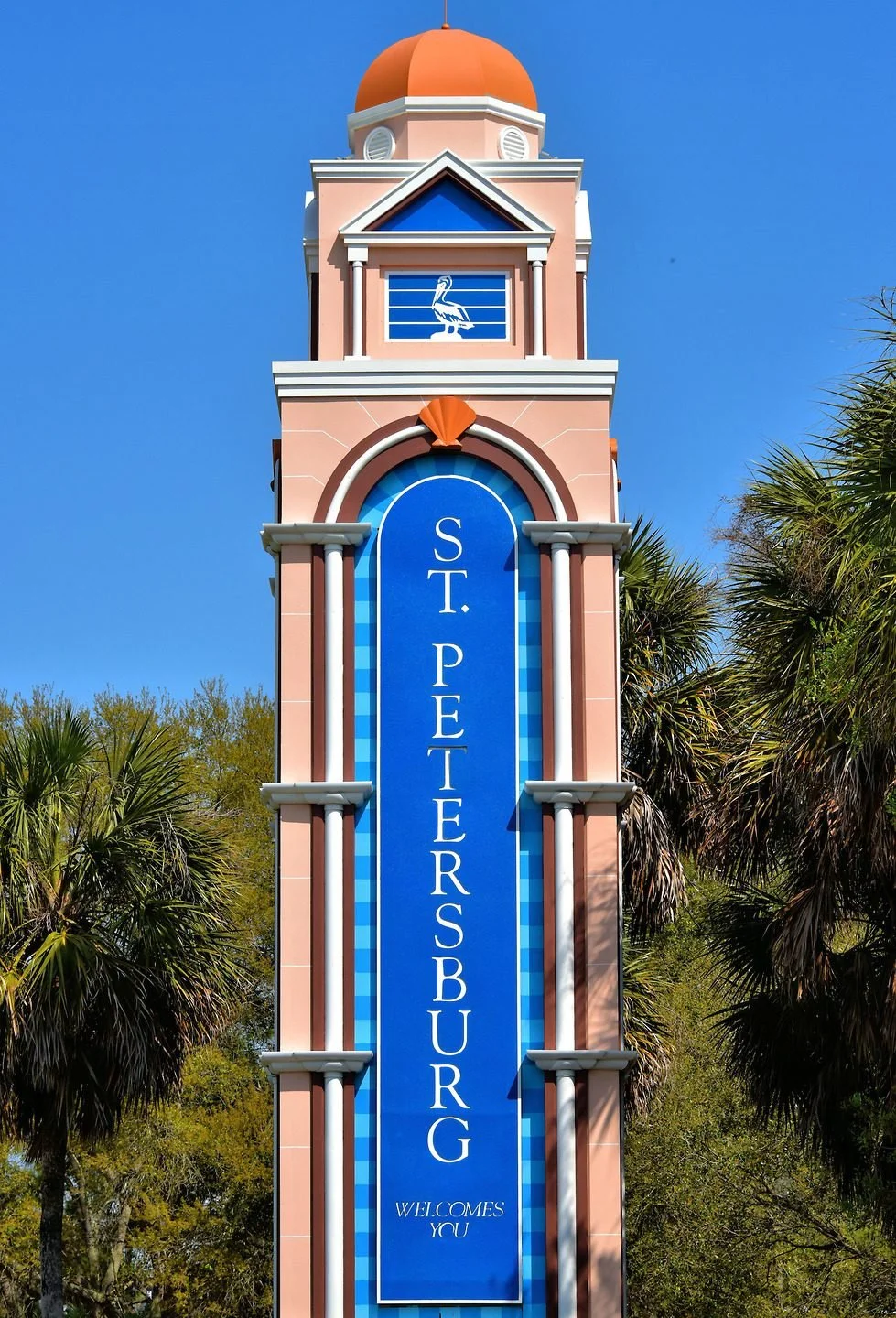 A clock tower with a pink and white exterior, topped with an orange domed roof, displaying a blue sign for St. Petersburg, Florida, with palm trees in the background against a clear blue sky.