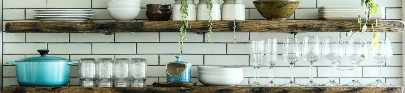 Kitchen shelves with dishes, glasses, bowls, mugs, and decorative items against white subway tile wall.