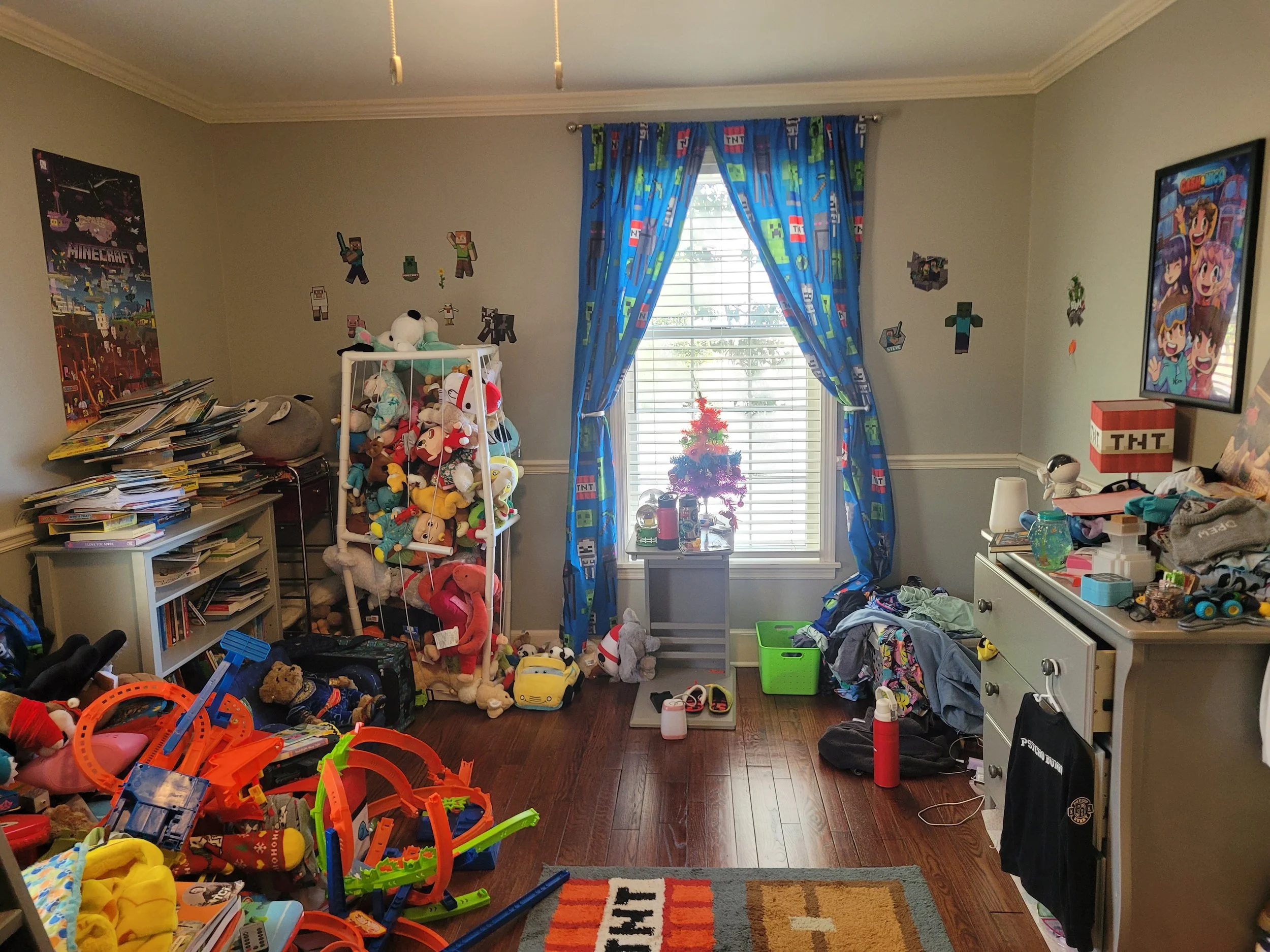 A cluttered children's bedroom with shelves of stuffed animals, toys, and books. Bright blue curtains with Minecraft characters cover a window, and posters decorate the walls. The floor has toy tracks, and various scattered toys and clothing fill the room.