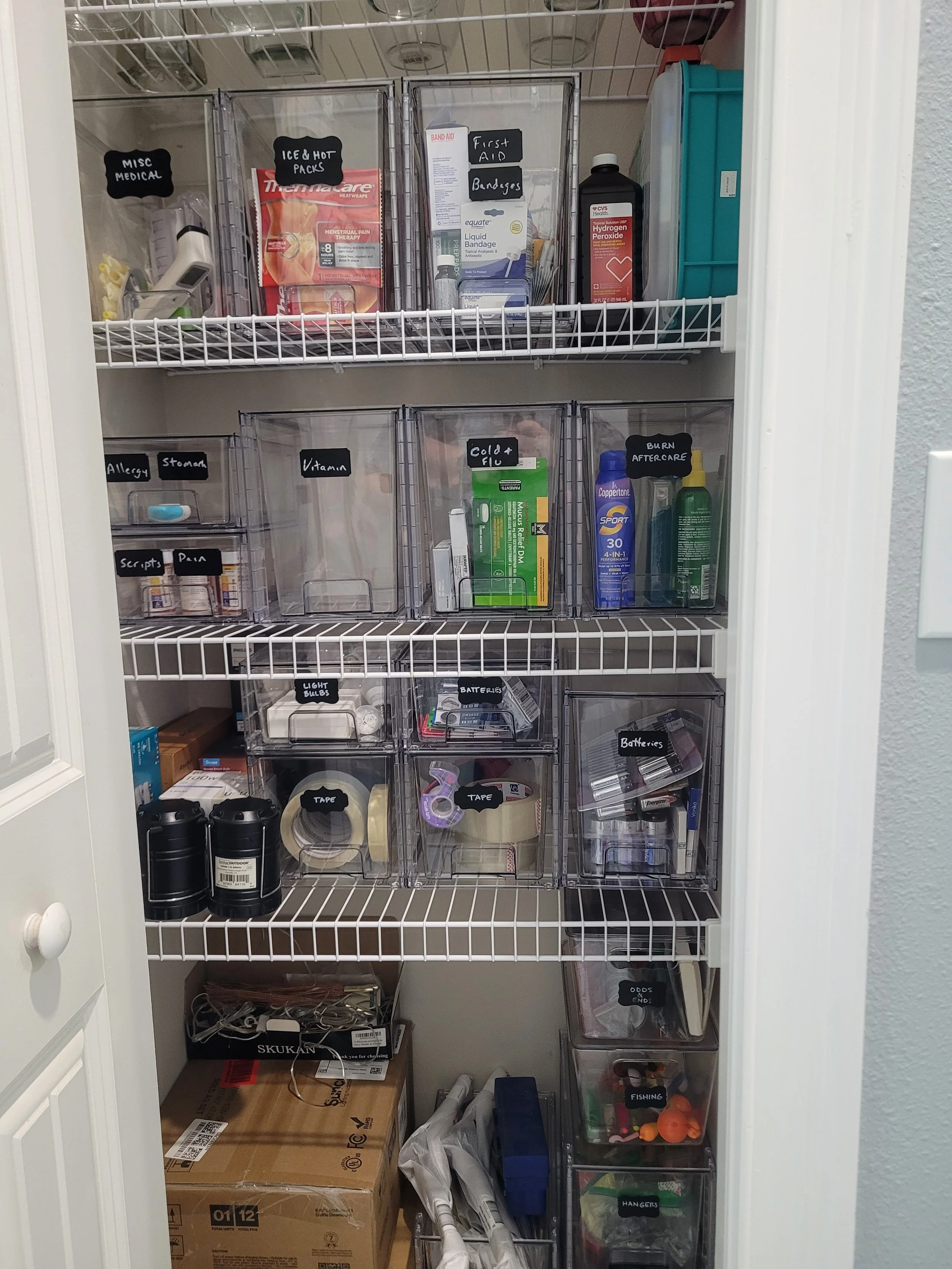 A storage closet with shelves containing various organized plastic bins labeled for medical supplies, cold and flu remedies, batteries, fishing gear, and other miscellaneous items. There are also boxes, bottles, and small supplies inside.