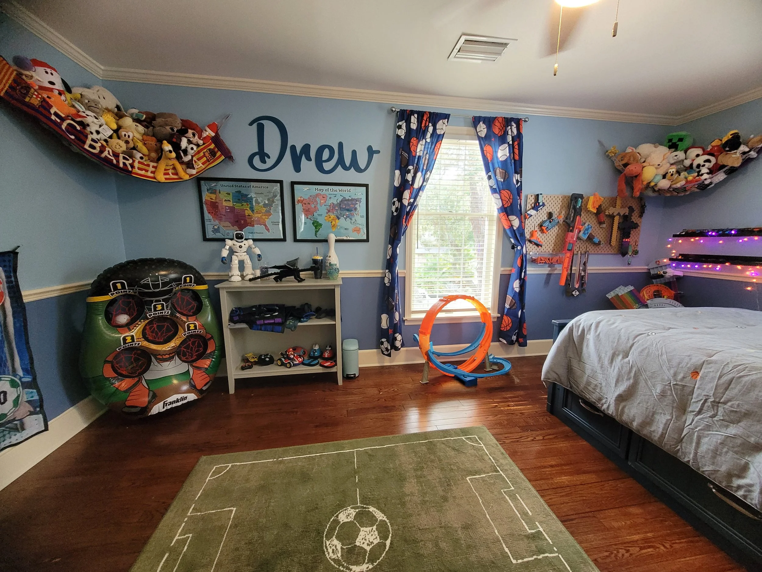 Children's bedroom with blue walls, sports-themed curtains, and toys including plush stuffed animals, toy cars, a dartboard, and a foam football target. There are two framed maps of the United States and the world on the wall, and a large green soccer ball-shaped plush toy on the floor.