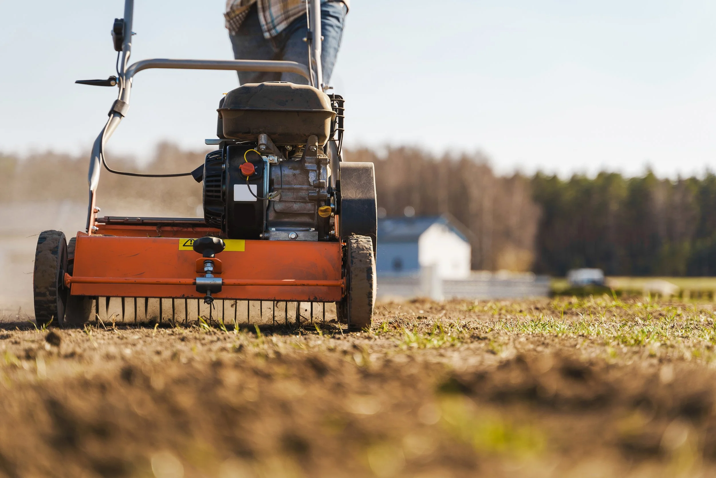 A person using a walk-behind rotary tiller on a farm field during daytime.