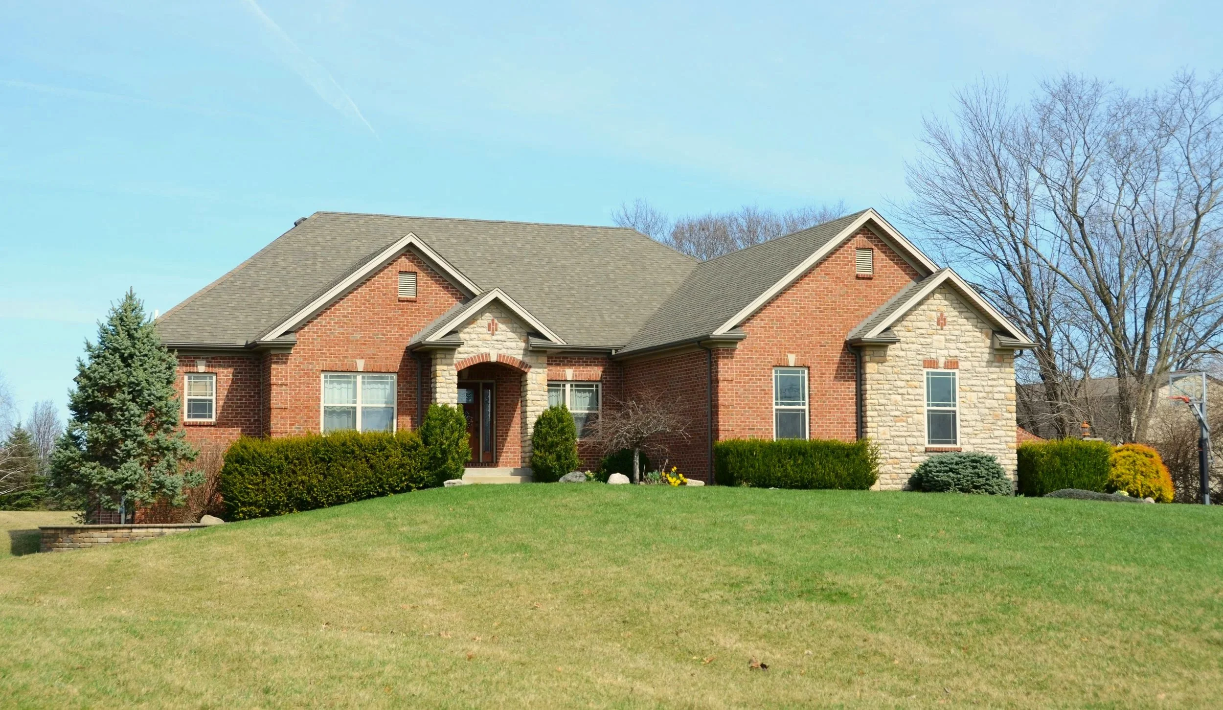 Front view of a large suburban house with brick and stone exterior, surrounded by trimmed bushes and trees in a green yard under a clear blue sky.
