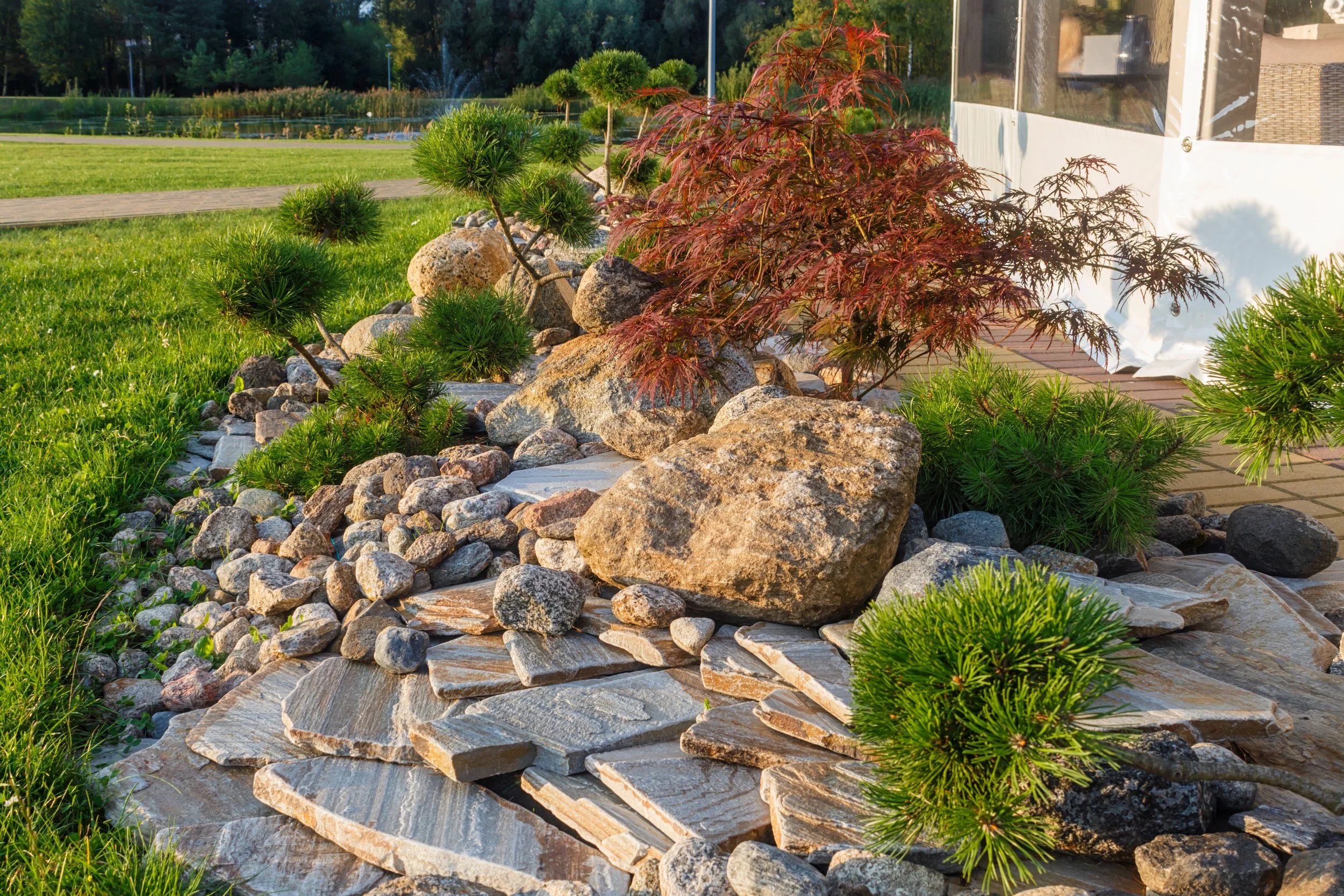 A landscaped garden with large rocks, small green shrubs, a red-leafed tree, and a grassy lawn in the background under a clear blue sky.