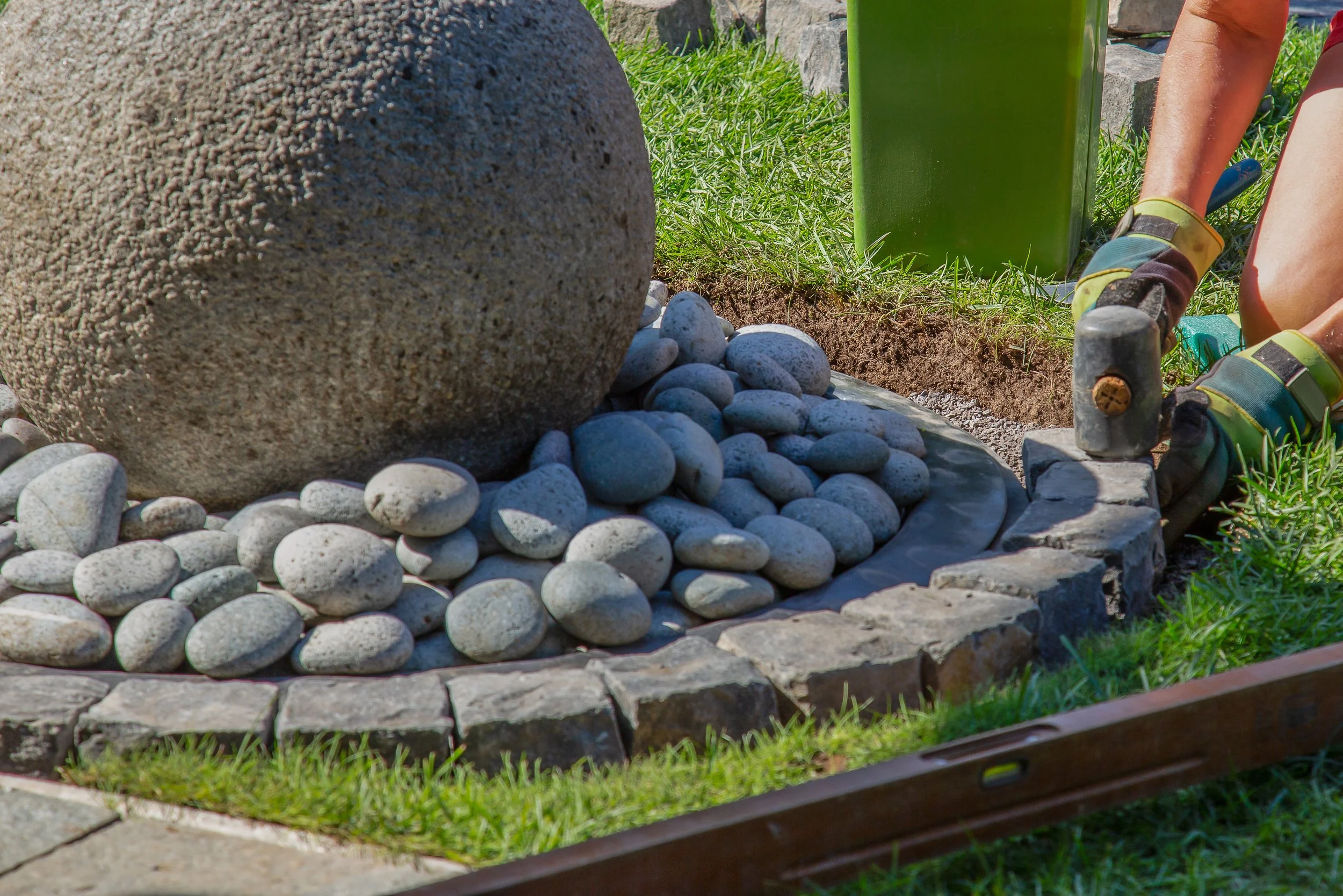 Person installing a stone border around a rock garden with a large boulder and small smooth gray stones, using a hammer and gloves.