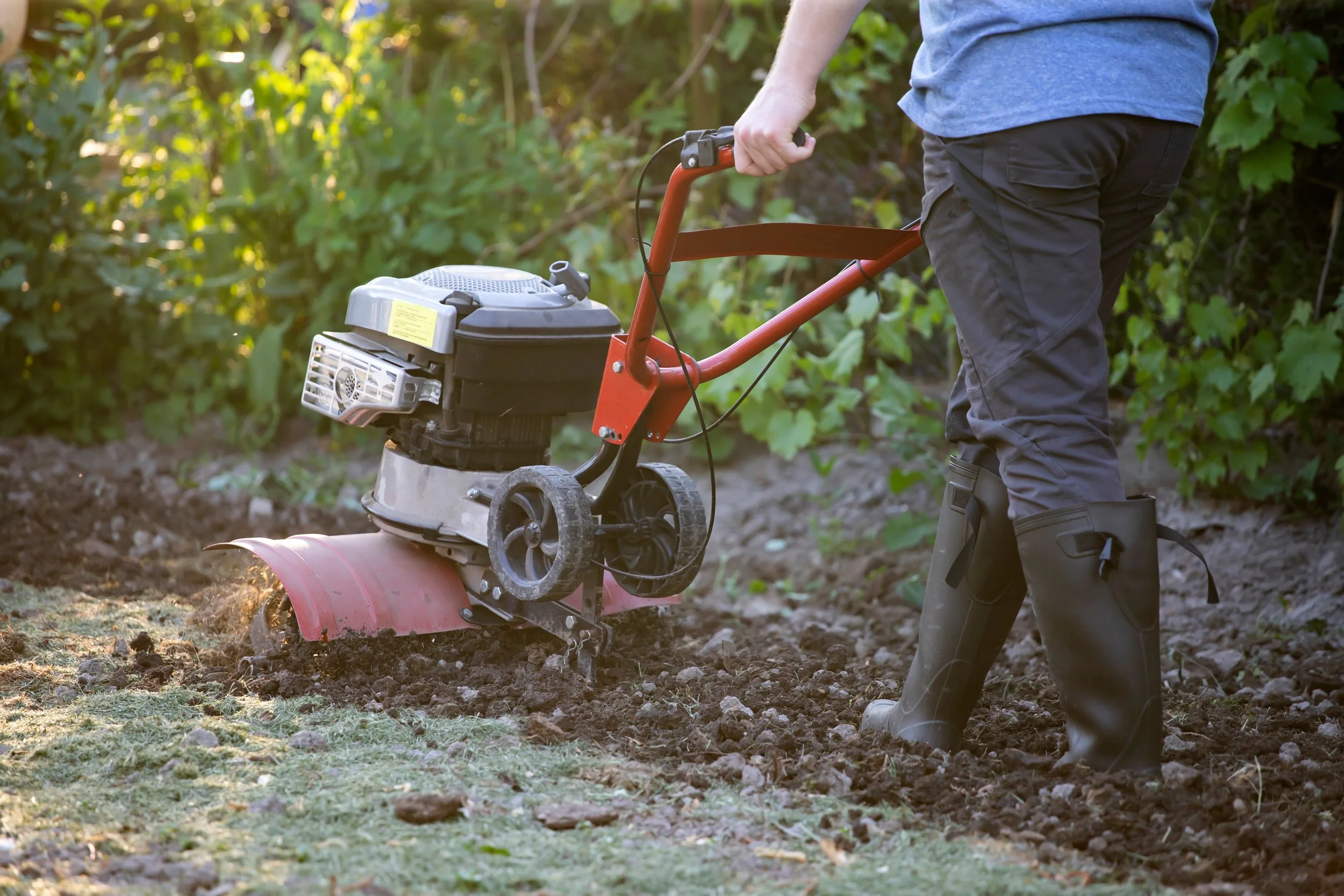 Person using a tiller in a garden against a backdrop of green foliage.