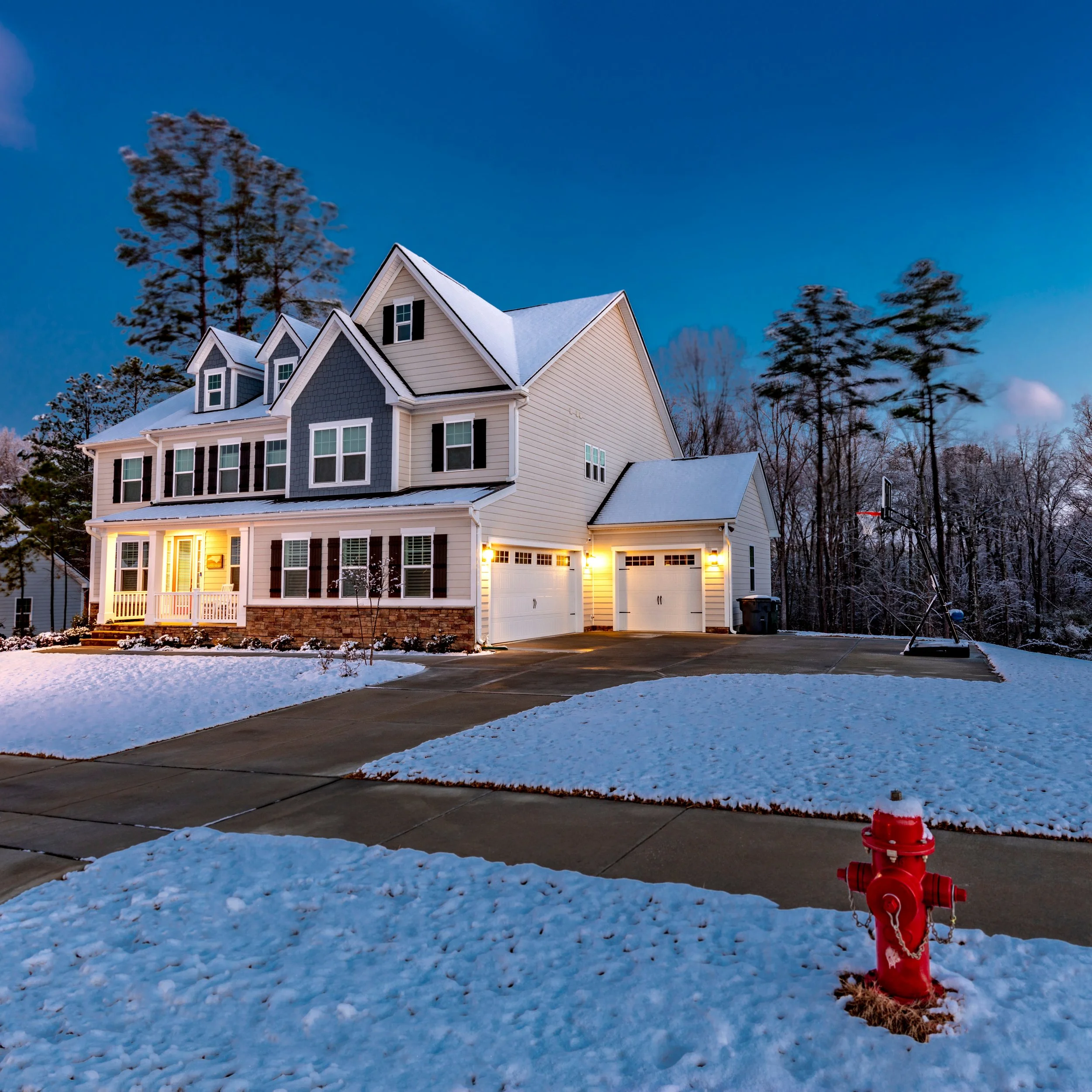 A large house with three stories, white siding, and black shutters, illuminated by exterior lights at dusk, surrounded by snow-covered lawn and trees, with a driveway, a basketball hoop, and a red fire hydrant.