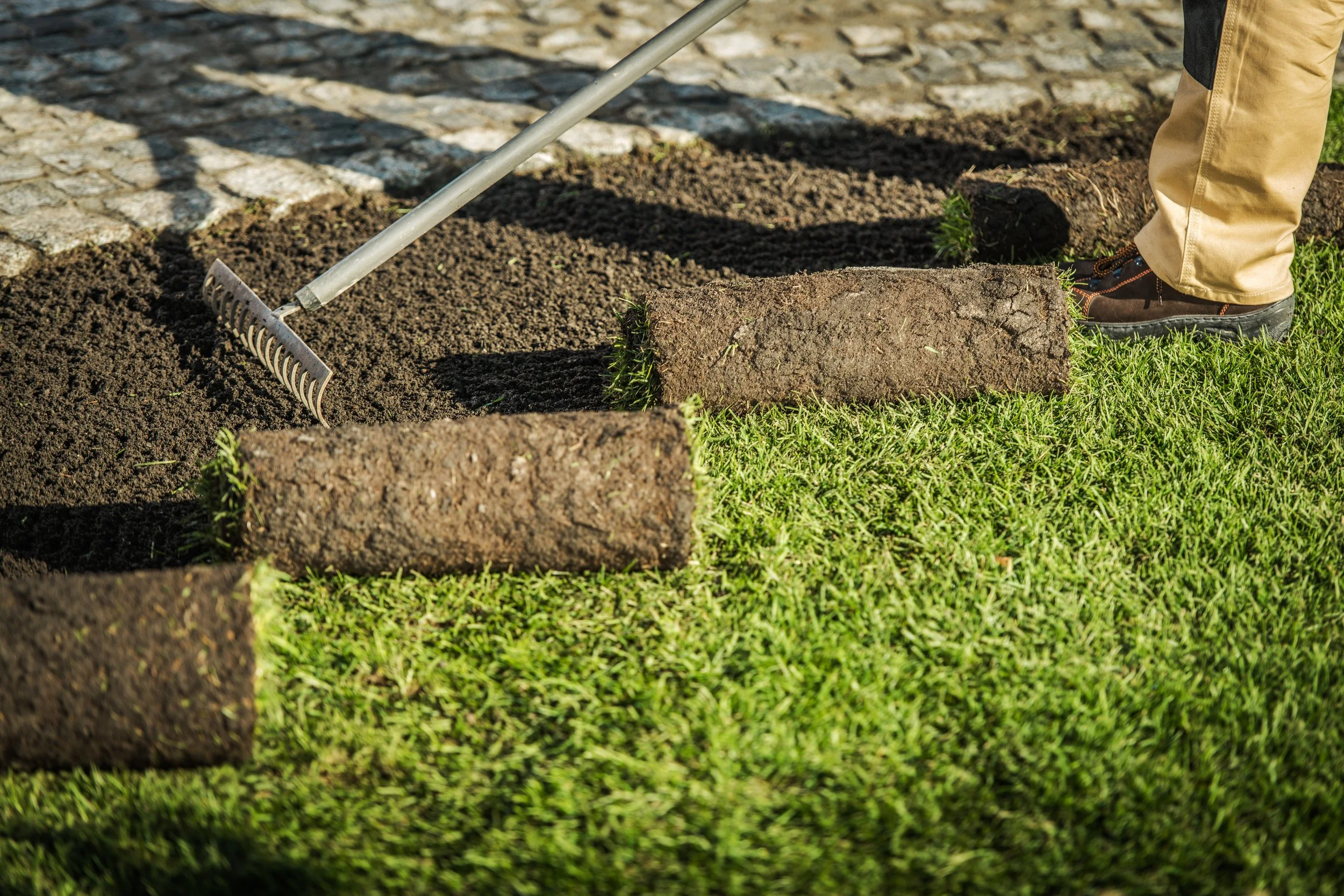 Person laying down bricks on a new garden bed with a rake, wearing beige pants and black shoes.