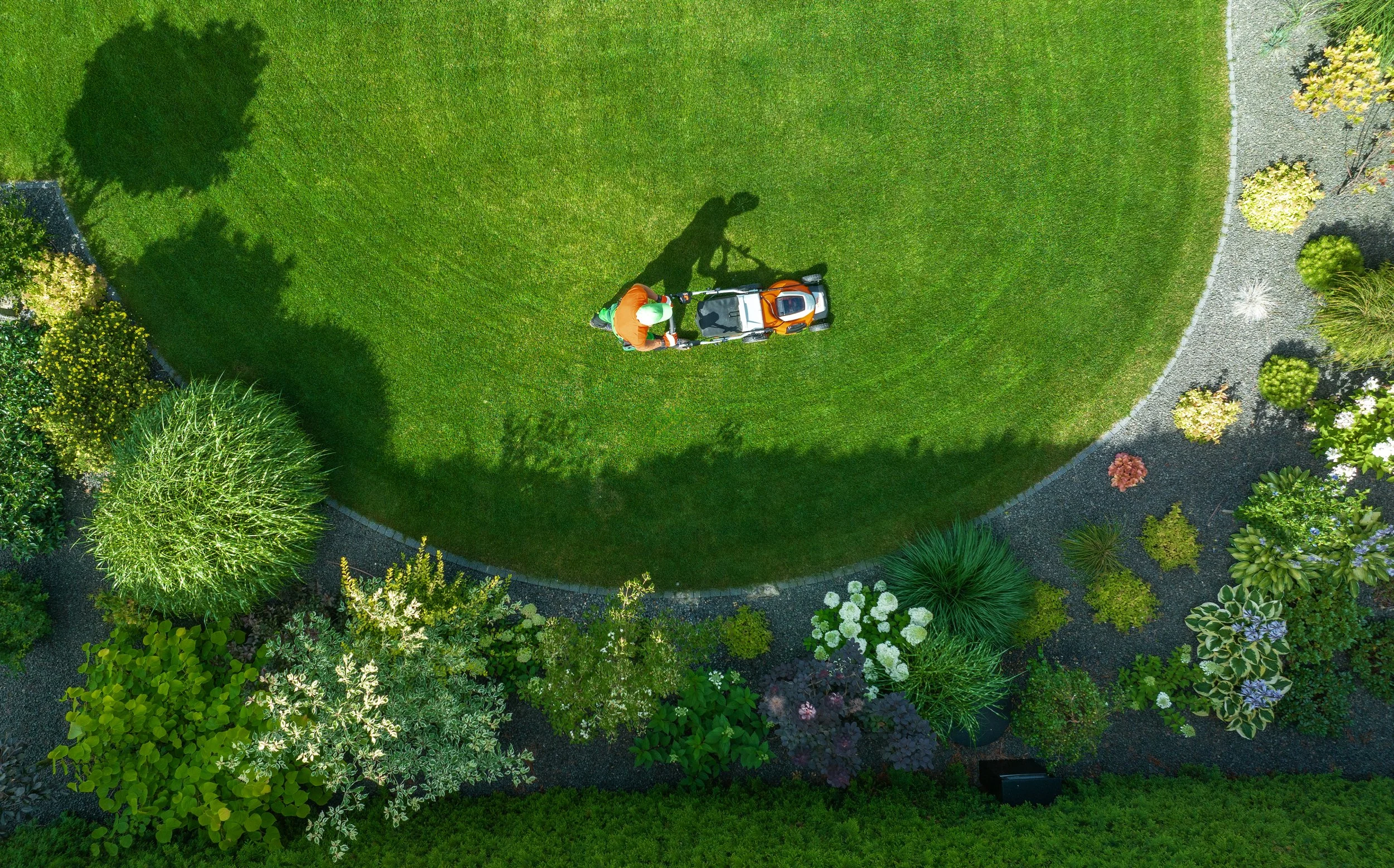 A person mowing a green lawn using a ride-on lawn mower in a landscaped garden with various plants and trees, viewed from above.
