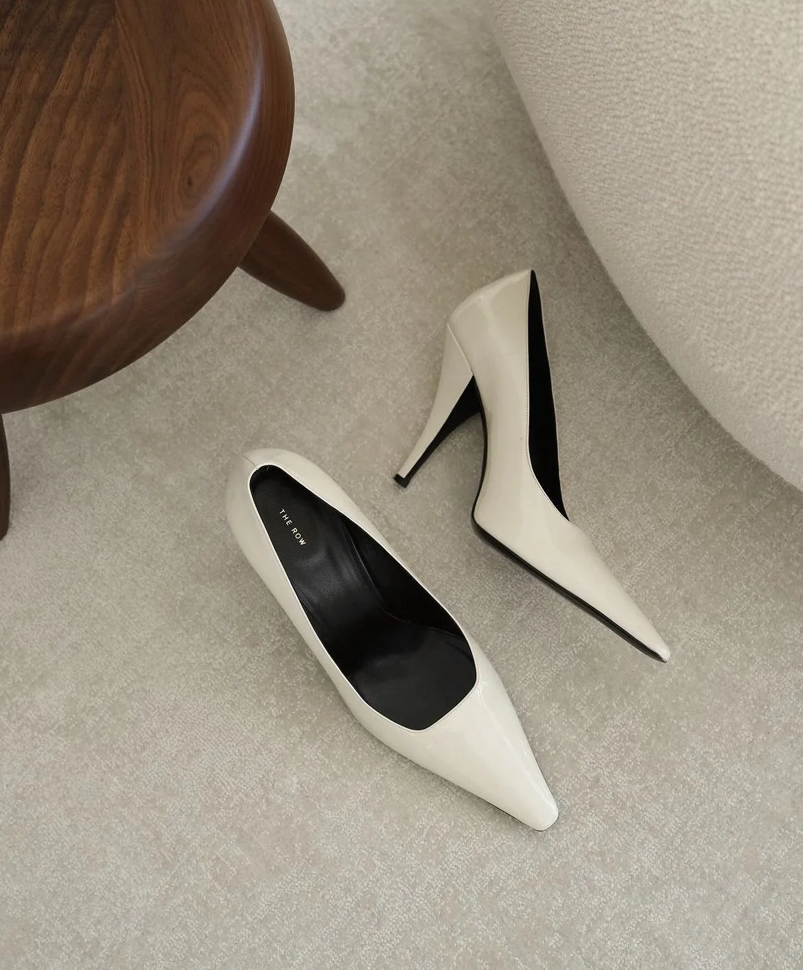 A pair of white high-heeled shoes with black insoles on light-colored carpet near a wooden table and a beige upholstered piece of furniture.