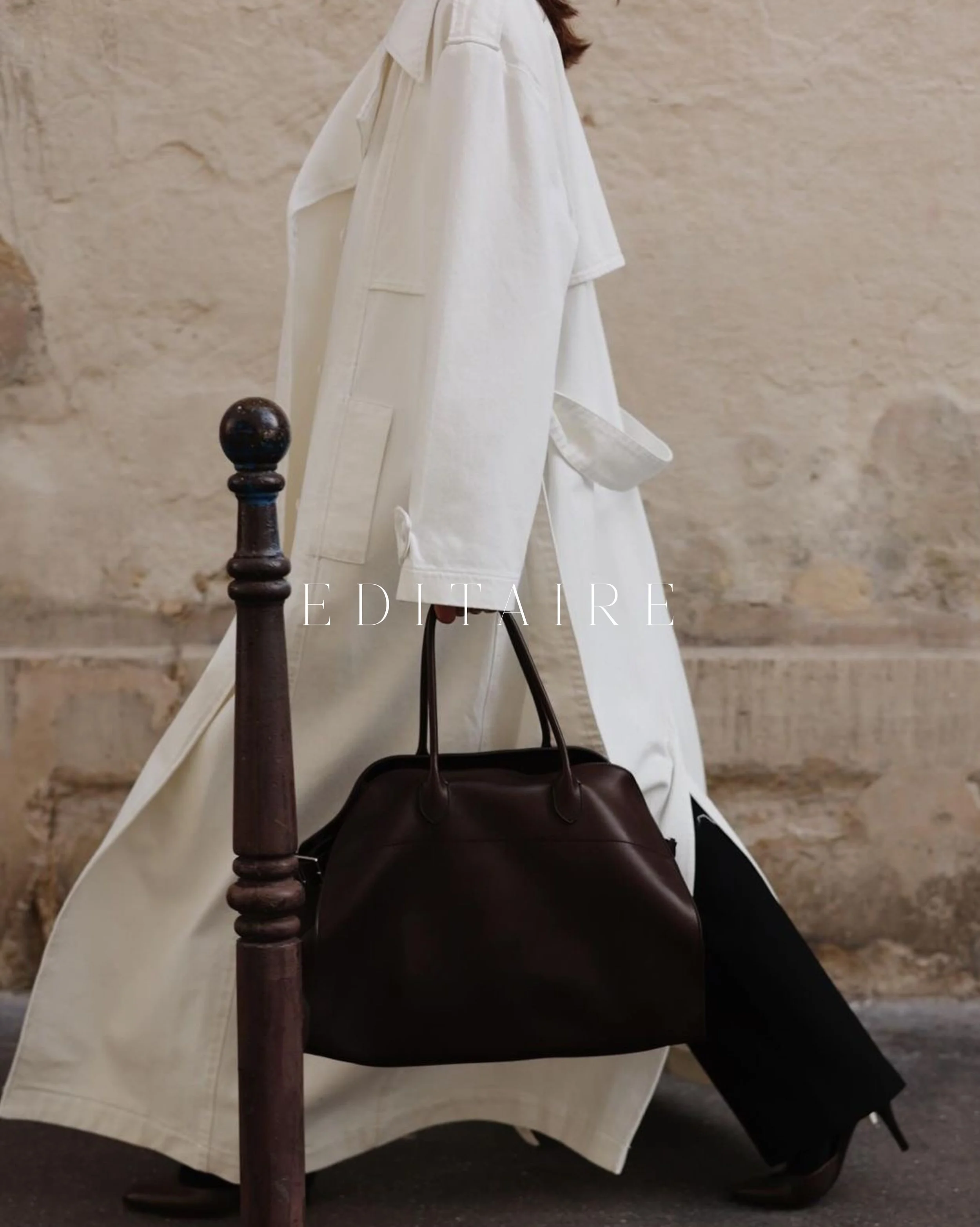 Person wearing a long white coat holding a dark brown leather handbag, standing next to a wooden police barricade against a textured beige wall.