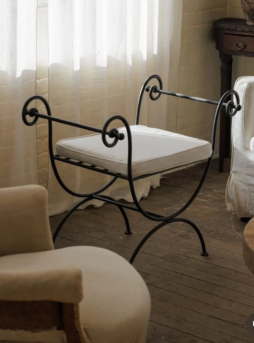 A vintage rocking cradle with a cushioned mattress, made of black metal with curved design, situated in a cozy room with wooden flooring, beige curtains, and surrounding furniture.