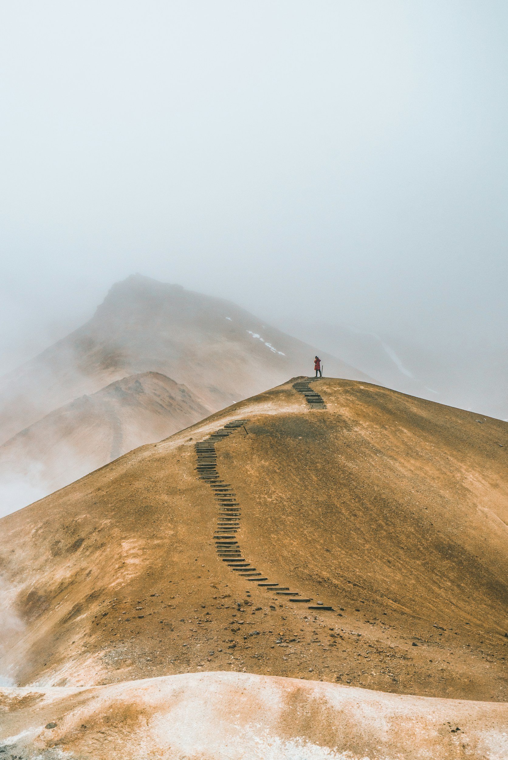 A person standing on a winding mountain path in a foggy landscape with barren, brown hills and distant mountains.