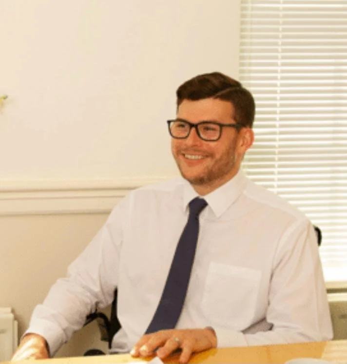 A man wearing glasses, a white shirt, and a dark tie, smiling while sitting at a desk in an office.