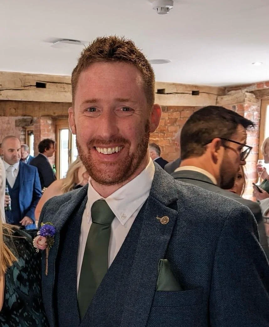 Smiling man with a beard in a suit at a social gathering in a rustic brick and wood interior.
