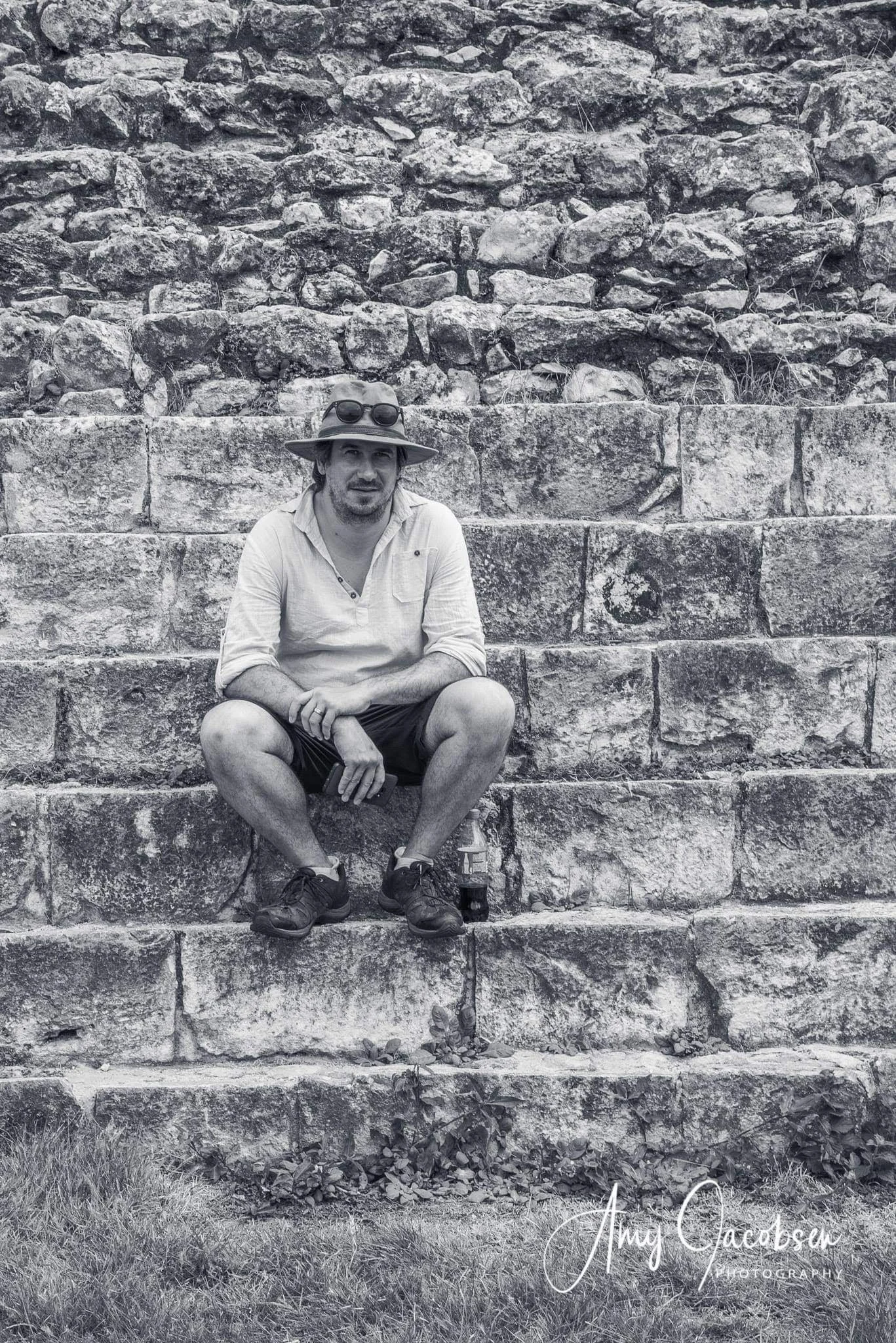 A man in a baseball cap with sunglasses on top, wearing a light shirt and shorts, sitting on stone steps against a stone wall, holding a bottle, with grass at the bottom of the image.