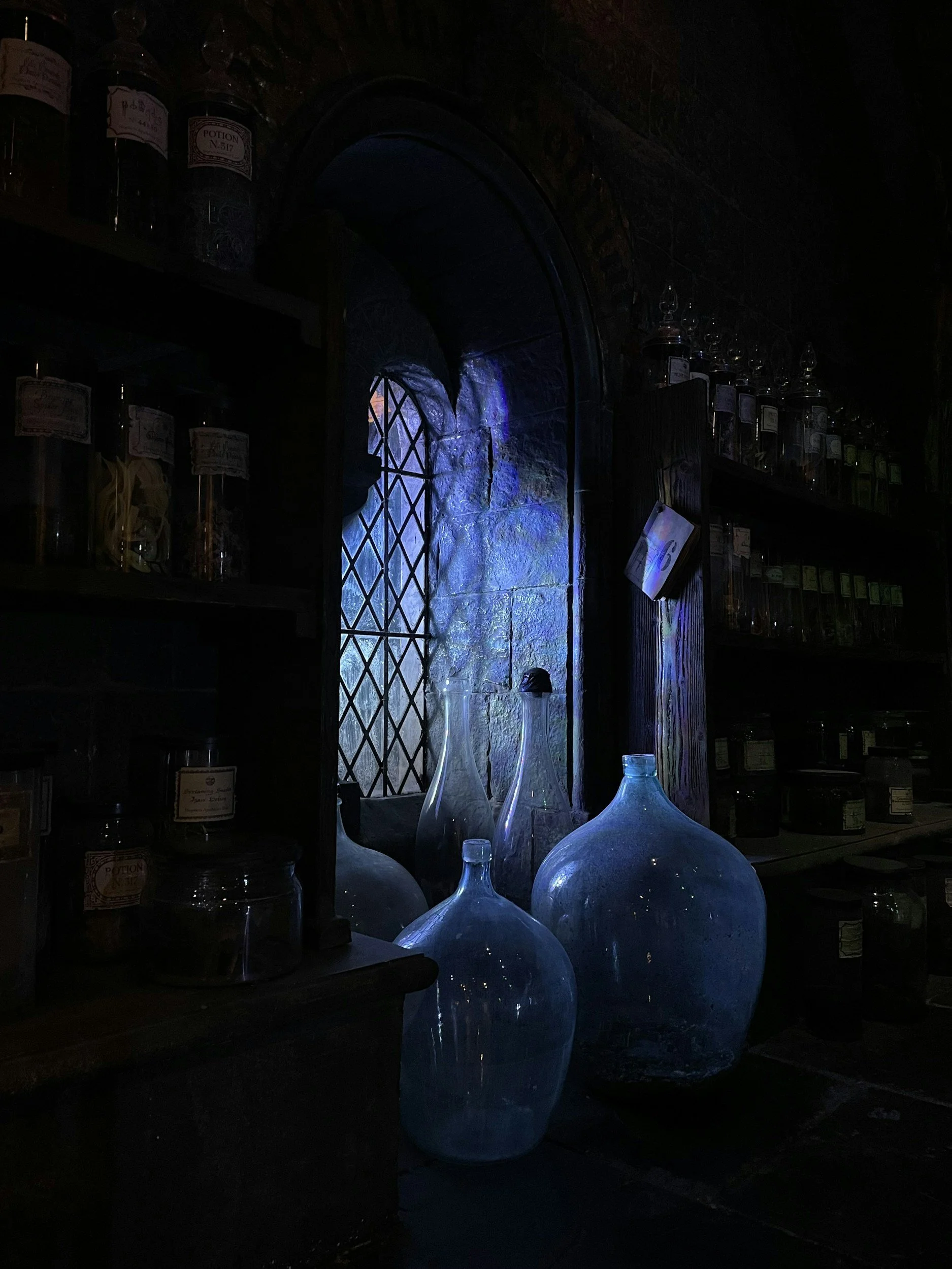 Dark interior with a small arched window and four large glass bottles reflecting blue light, shelves with jars and bottles, and an old wooden counter.