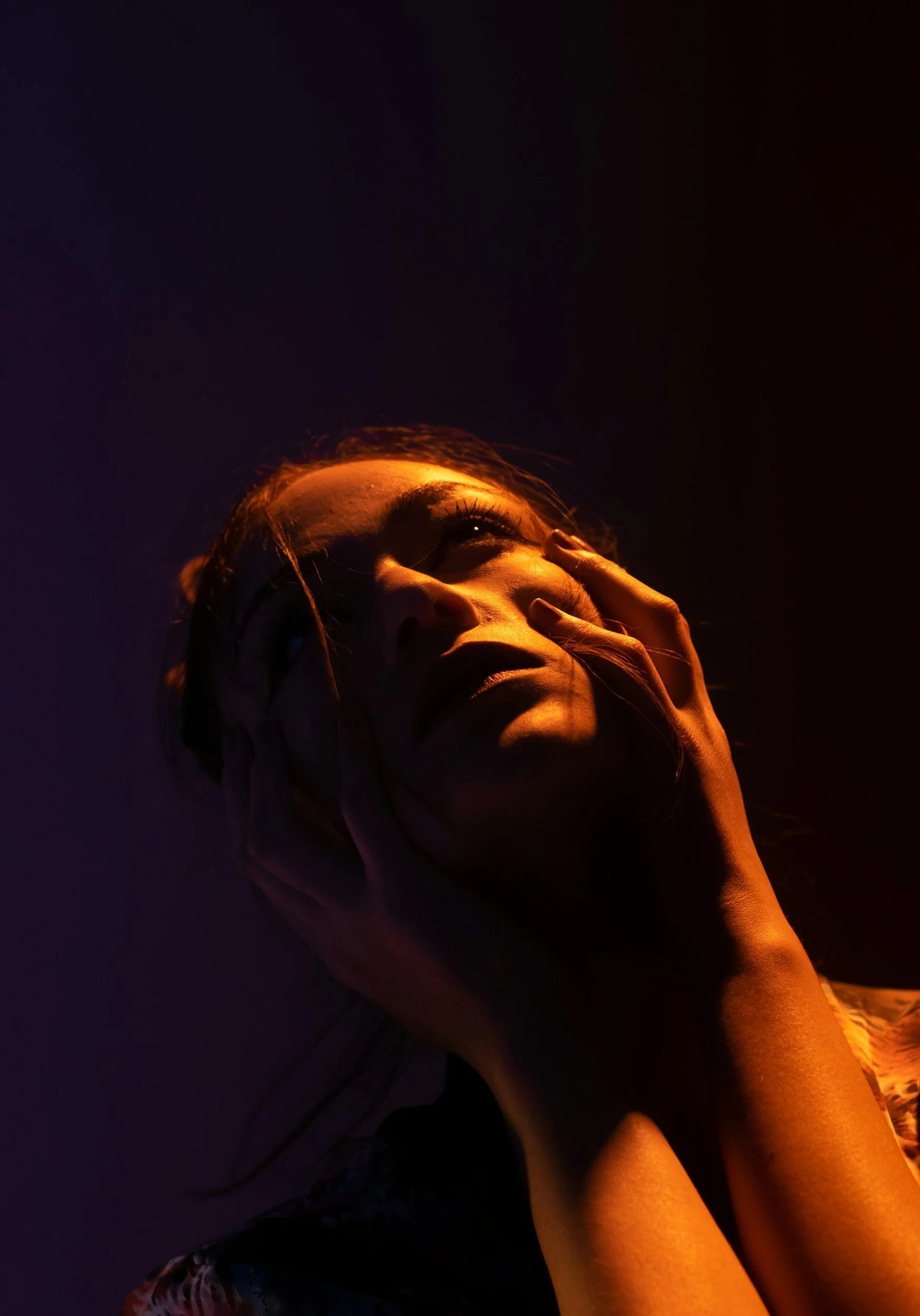 A woman with her hand on her face, illuminated by dramatic warm and cool lighting against a dark background.