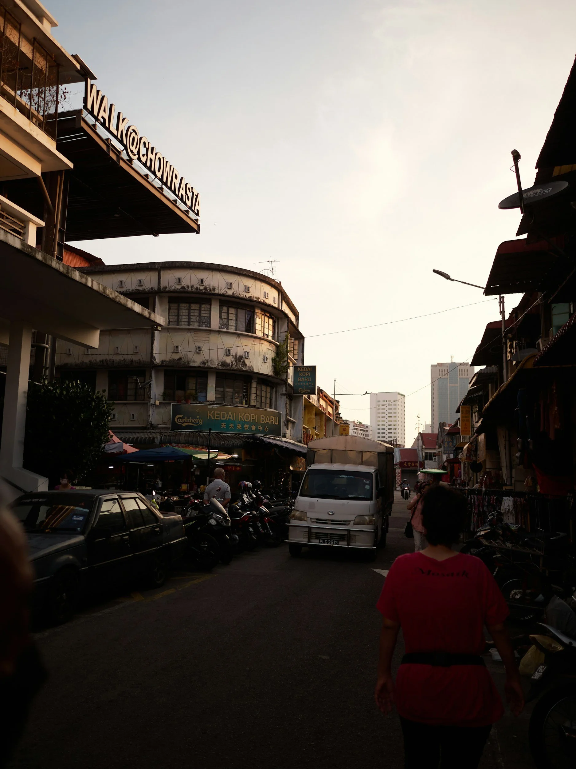 City street scene with buildings, parked motorcycles, cars, and a woman walking, during sunset or late afternoon.