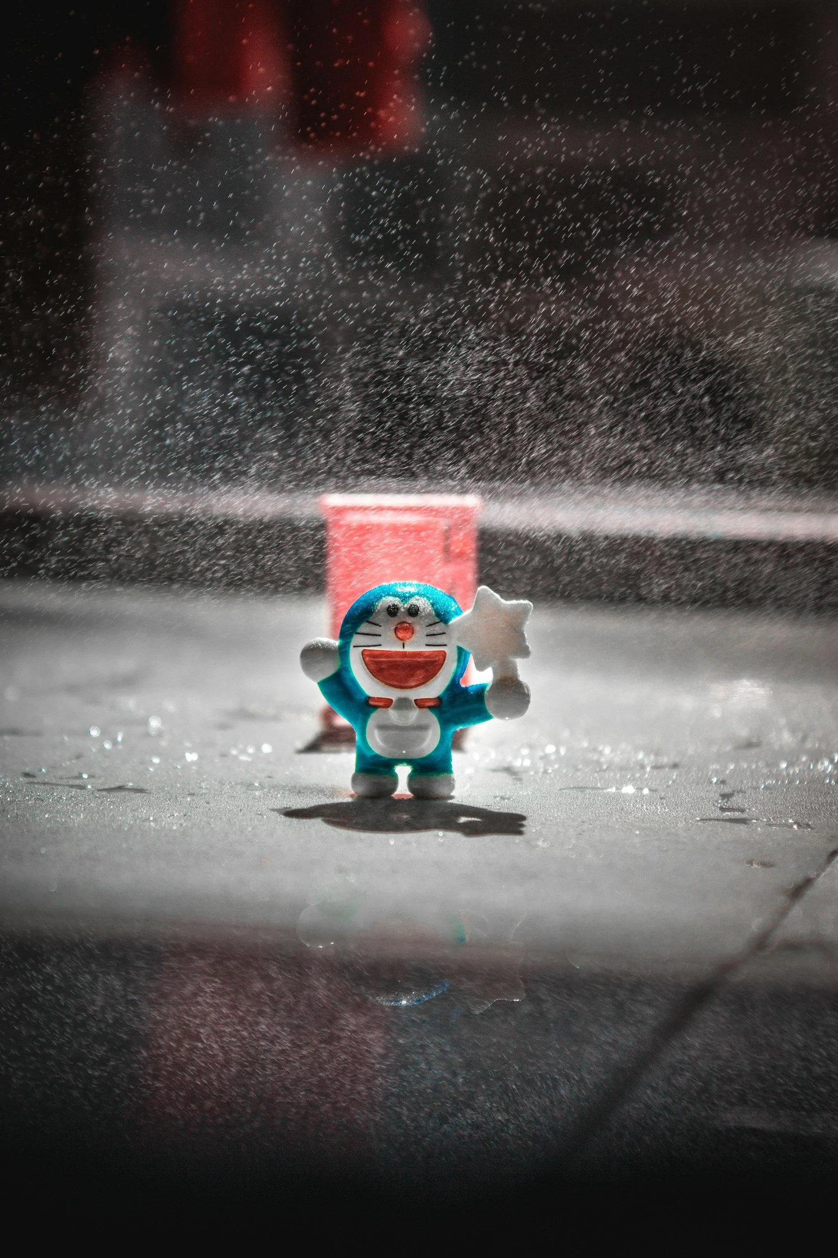 A small plush toy of Doraemon, a Japanese cartoon character, standing on a surface with snow or frost, with a small pink container and snow falling around it.