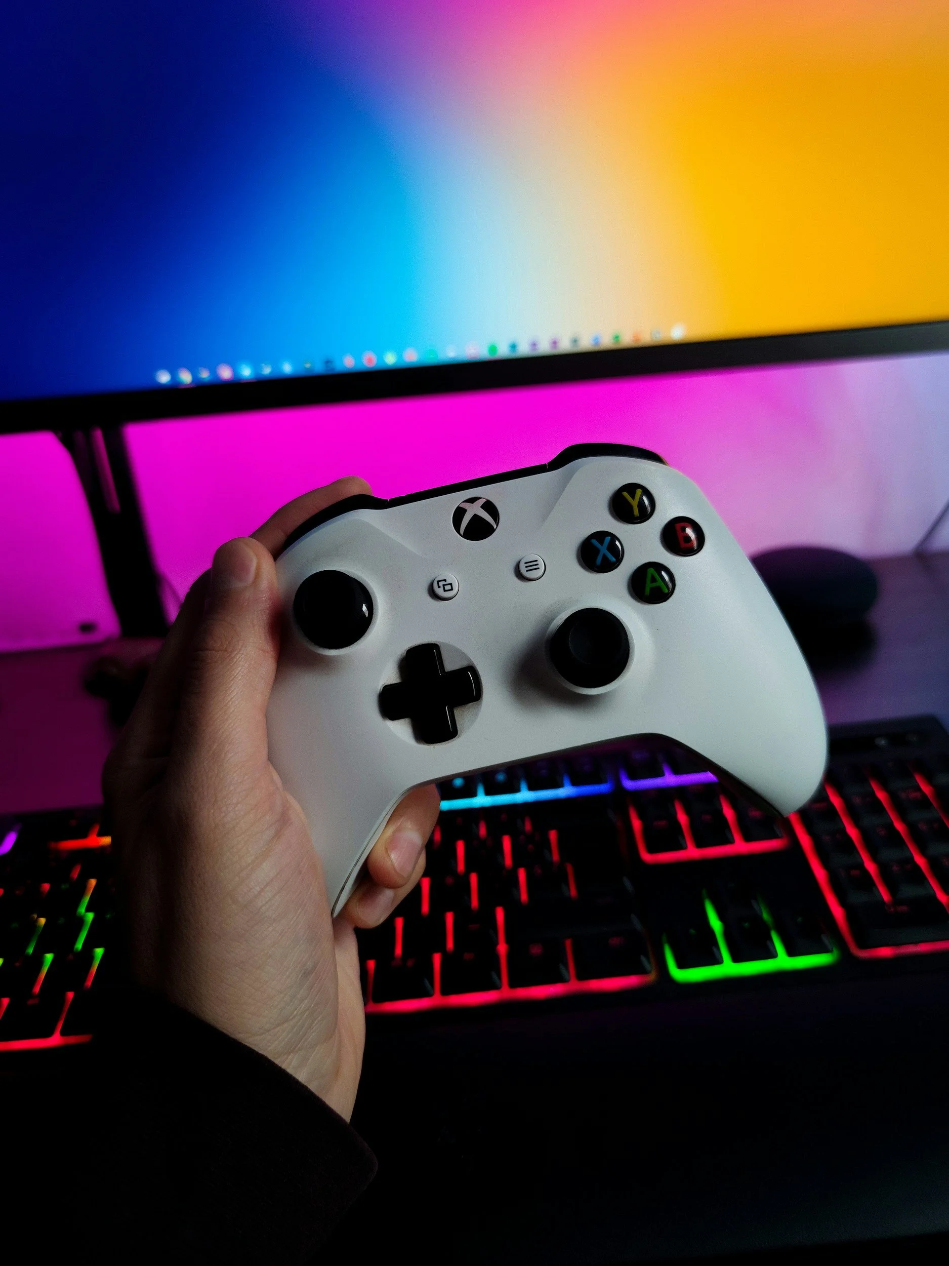 A person holding a white Xbox controller in front of a gaming setup with a colorful lit keyboard, a large monitor displaying a gradient background from blue to orange, and a mouse on the desk.