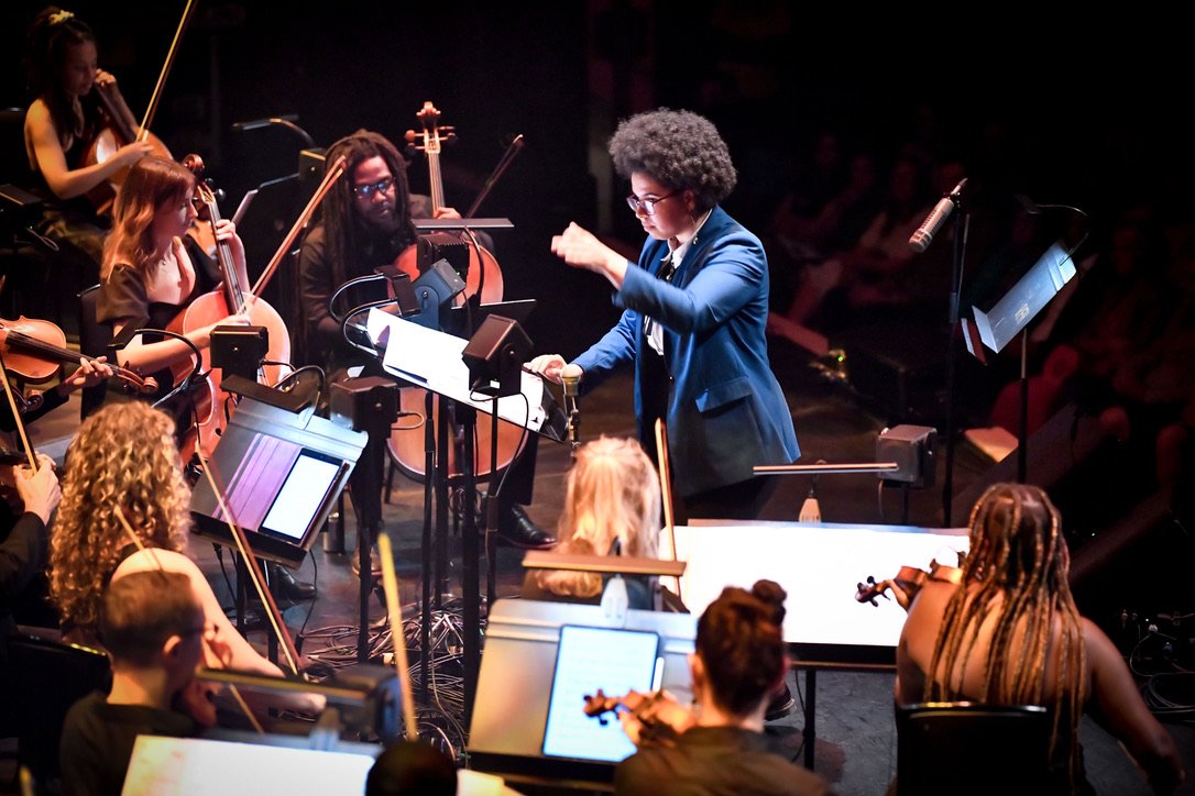 An orchestra group performing on stage, conducted by a person with curly hair wearing glasses and a blue jacket, with musicians playing string instruments.