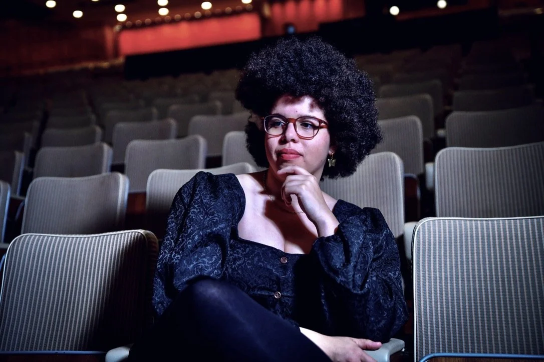 A woman with curly black hair and glasses sitting alone in an empty theater, resting her chin on her hand, looking thoughtful.