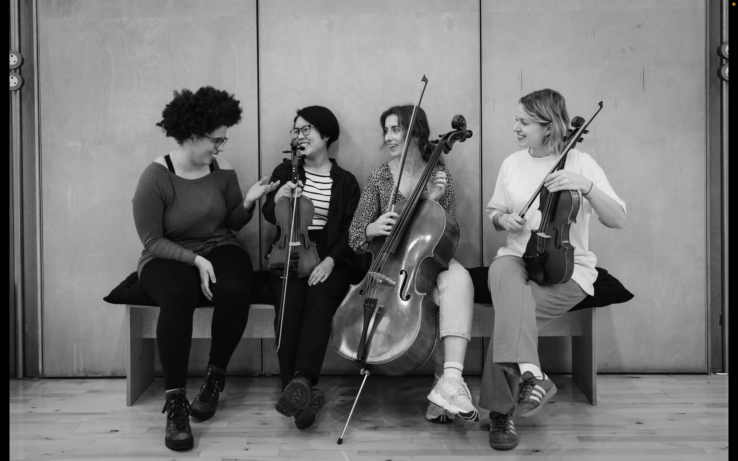 Four women sitting on a bench, engaged in conversation, with three playing musical instruments: a violin, a cello, and a viola.
