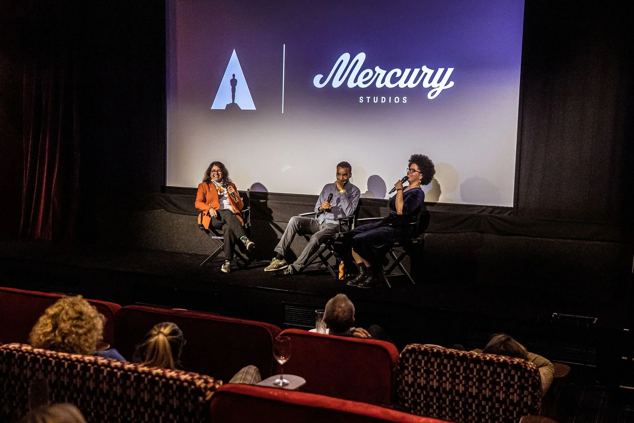 Panel discussion at a movie theater with three people on stage, one woman and two men, seated in chairs and talking into microphones, with a large screen behind them displaying the Mercury Studios logo.