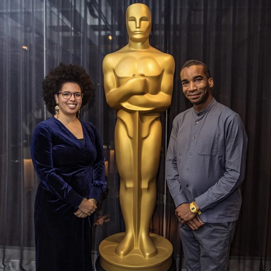 Two people, a woman and a man, standing on either side of a large gold award statue resembling an Oscar, inside a room with dark curtains in the background.