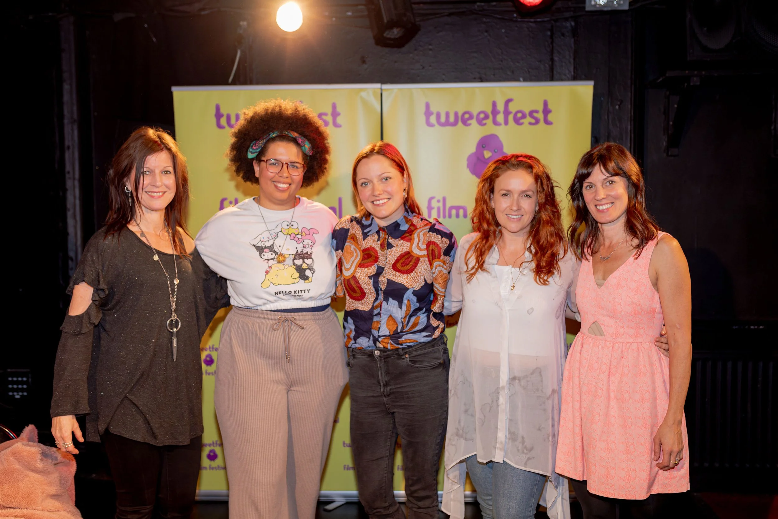 Five women standing together on a stage with purple and yellow 'tweetchest' banners in the background, smiling at the camera.