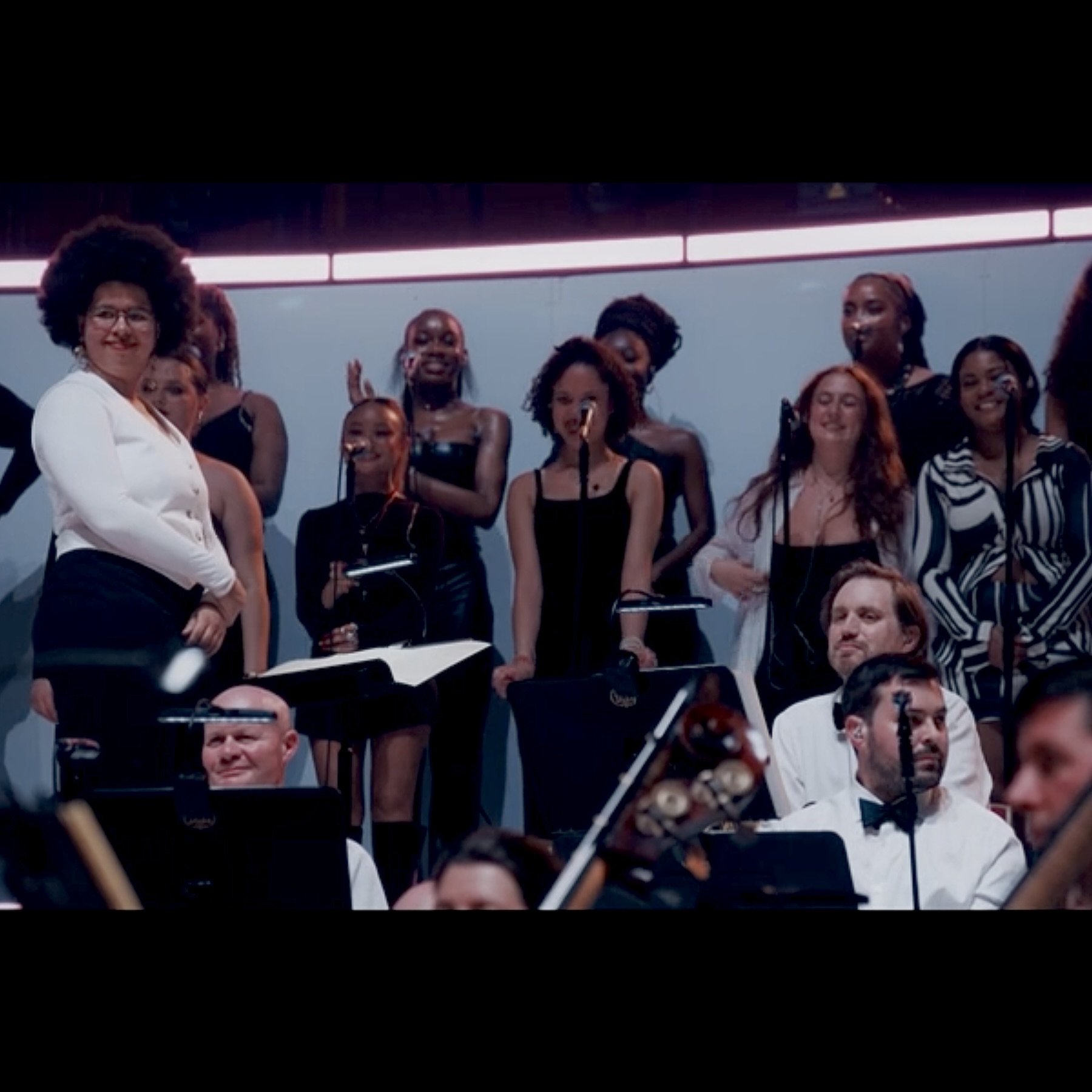 A diverse group of singers and musicians performing on stage, with some holding sheet music, in an orchestral concert setting.