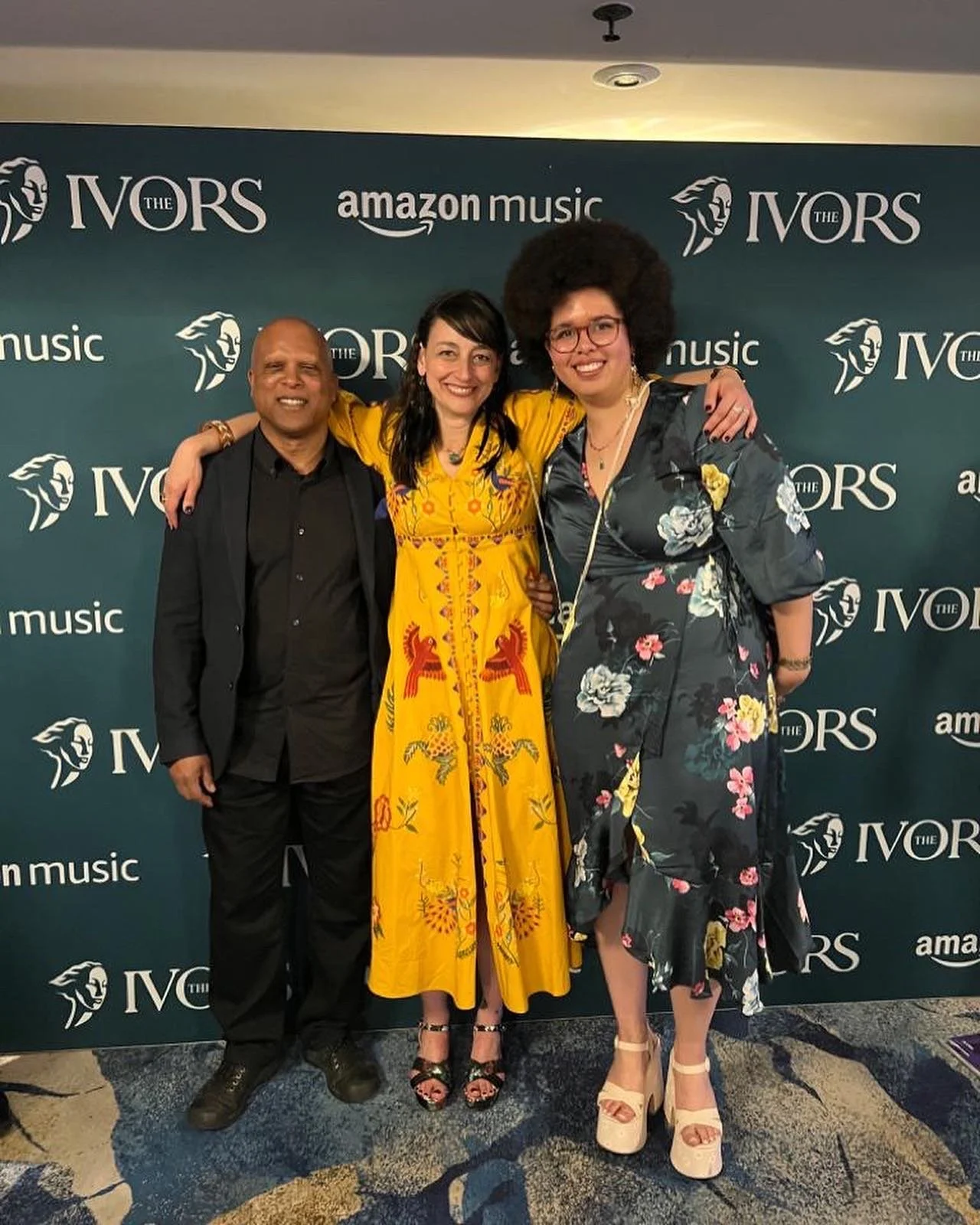 Three people posing together in front of a backdrop with 'The Ivors' and 'Amazon Music' logos. Two women and one man, all dressed in formal attire, smiling and with arms around each other.