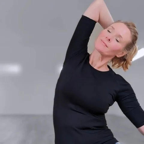 A woman in a black shirt smiling with her eyes closed, stretching her arm behind her head.