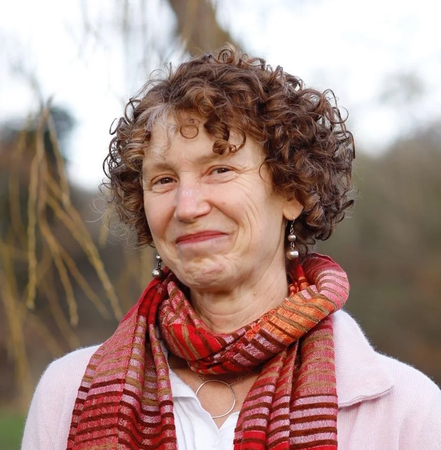 A woman with curly brown hair smiling outdoors, wearing a white top, a red patterned scarf, and earrings.