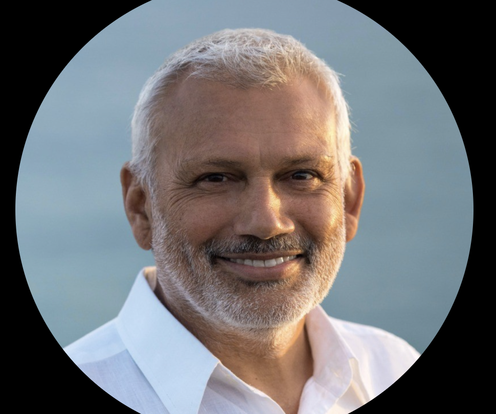 Smiling middle-aged man with gray hair and beard wearing a white collared shirt, outdoors with a blue sky background.
