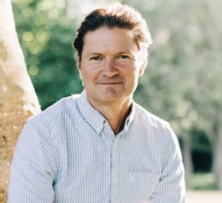 A man with dark hair and a light striped shirt standing outdoors with trees in the background.