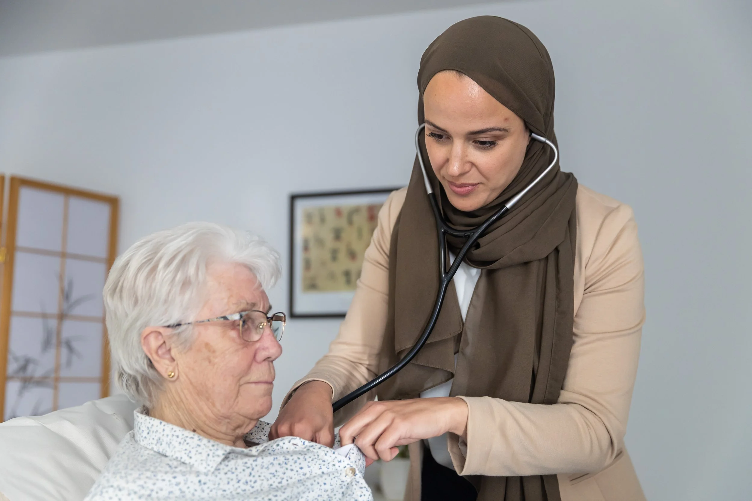 A healthcare professional wearing a hijab uses a stethoscope to check an elderly woman's heartbeat in a medical setting.