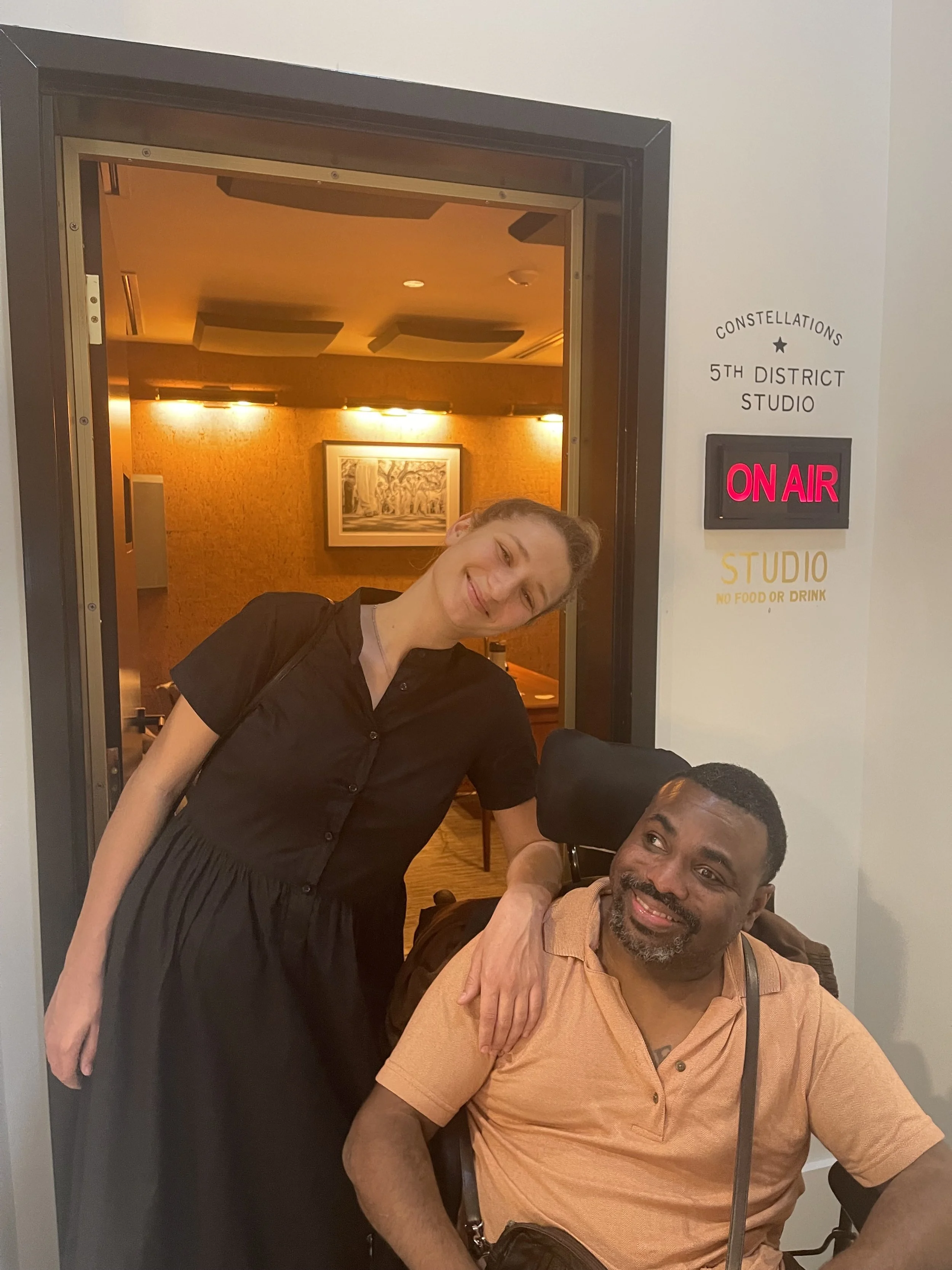 Two smiling people, a woman and a man in a wheelchair, inside a studio with a sign indicating 'On Air'. The woman is standing with her arm on the man's shoulder, leaning her head towards him.