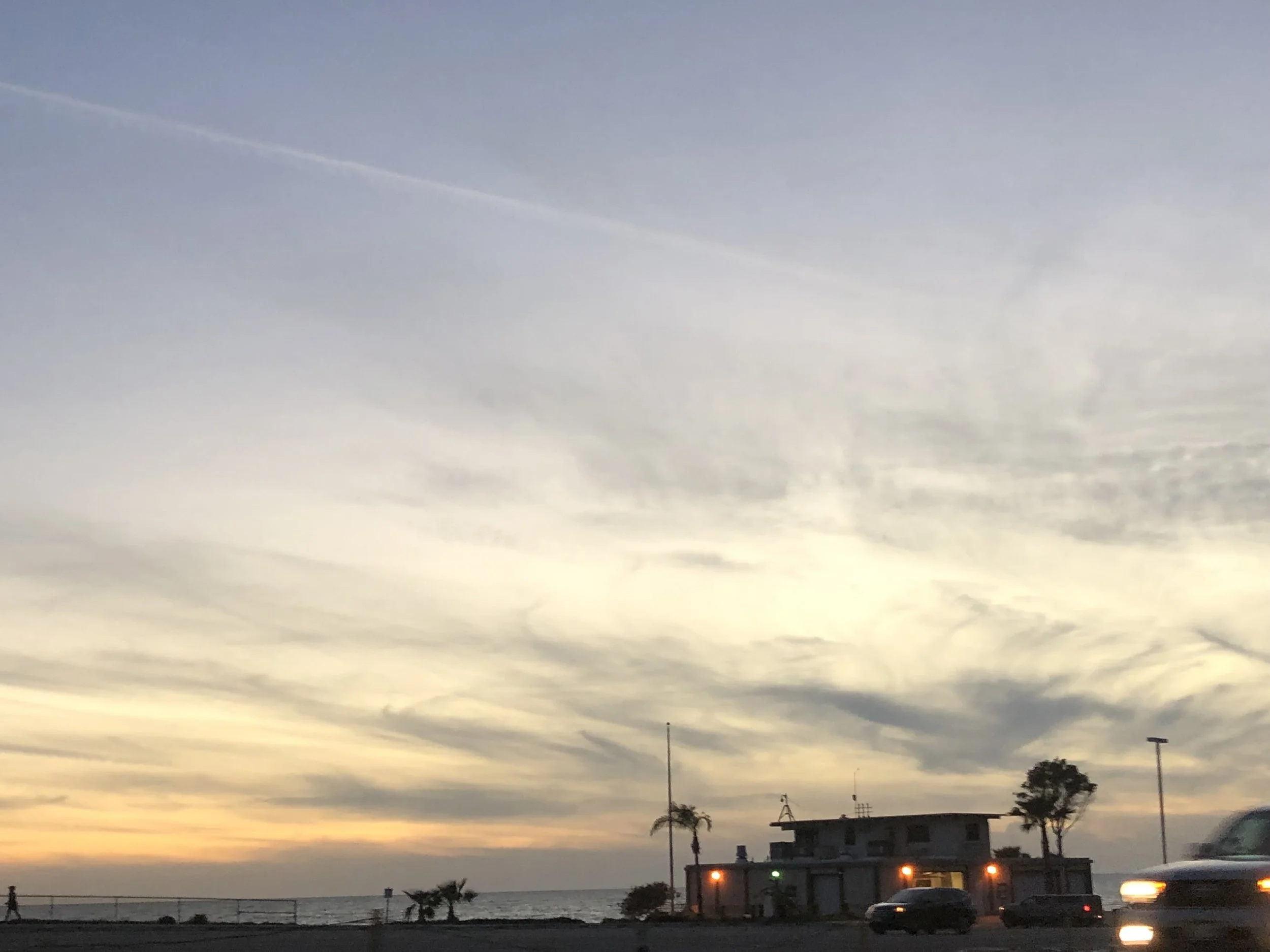Sunset by the seaside with a cloudy sky, a building, a few palm trees, and cars on the road.