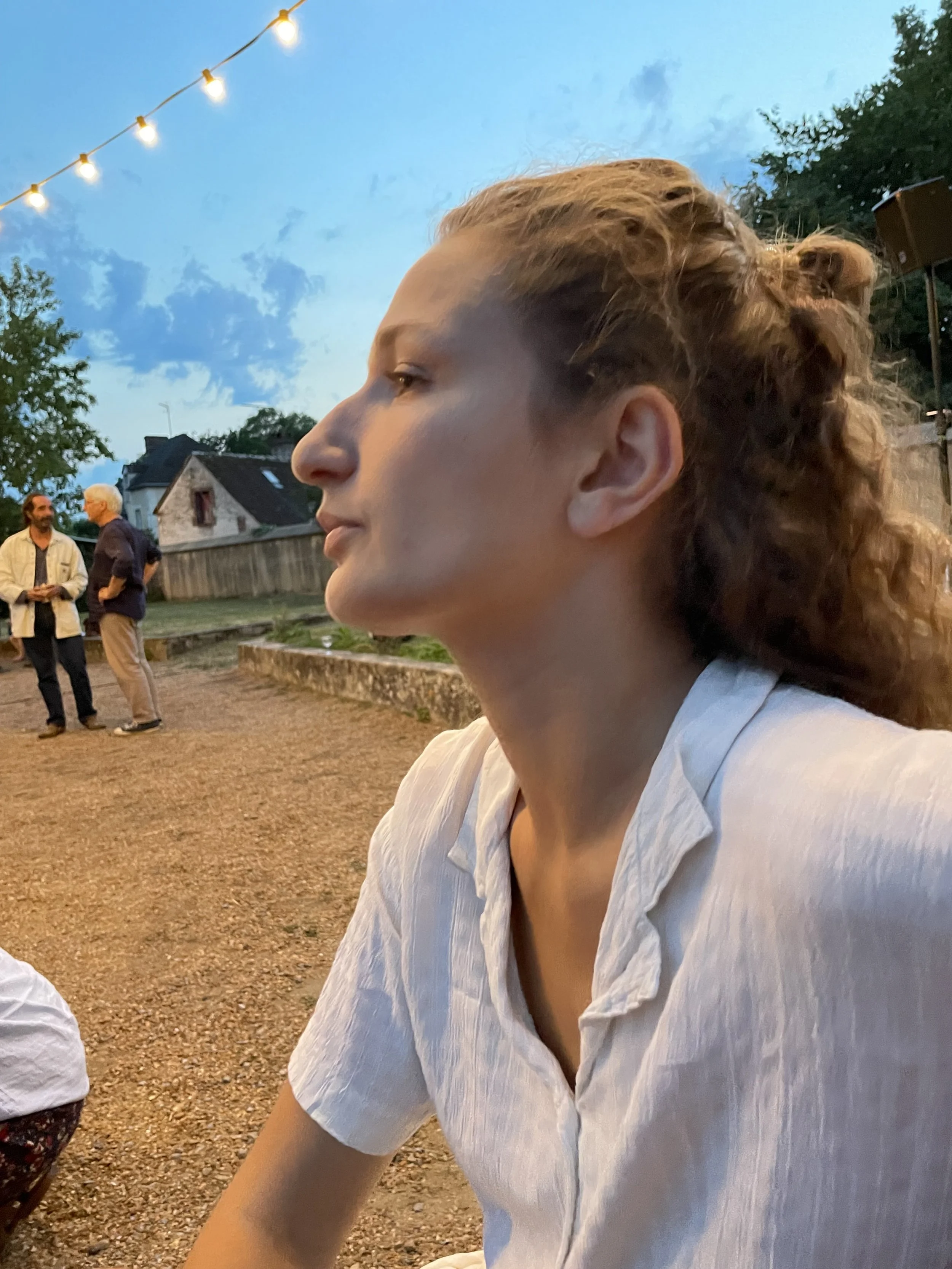 A woman with curly hair tied back, wearing a light blue shirt, is sitting outside during an evening, with string lights above her and people talking in the background.