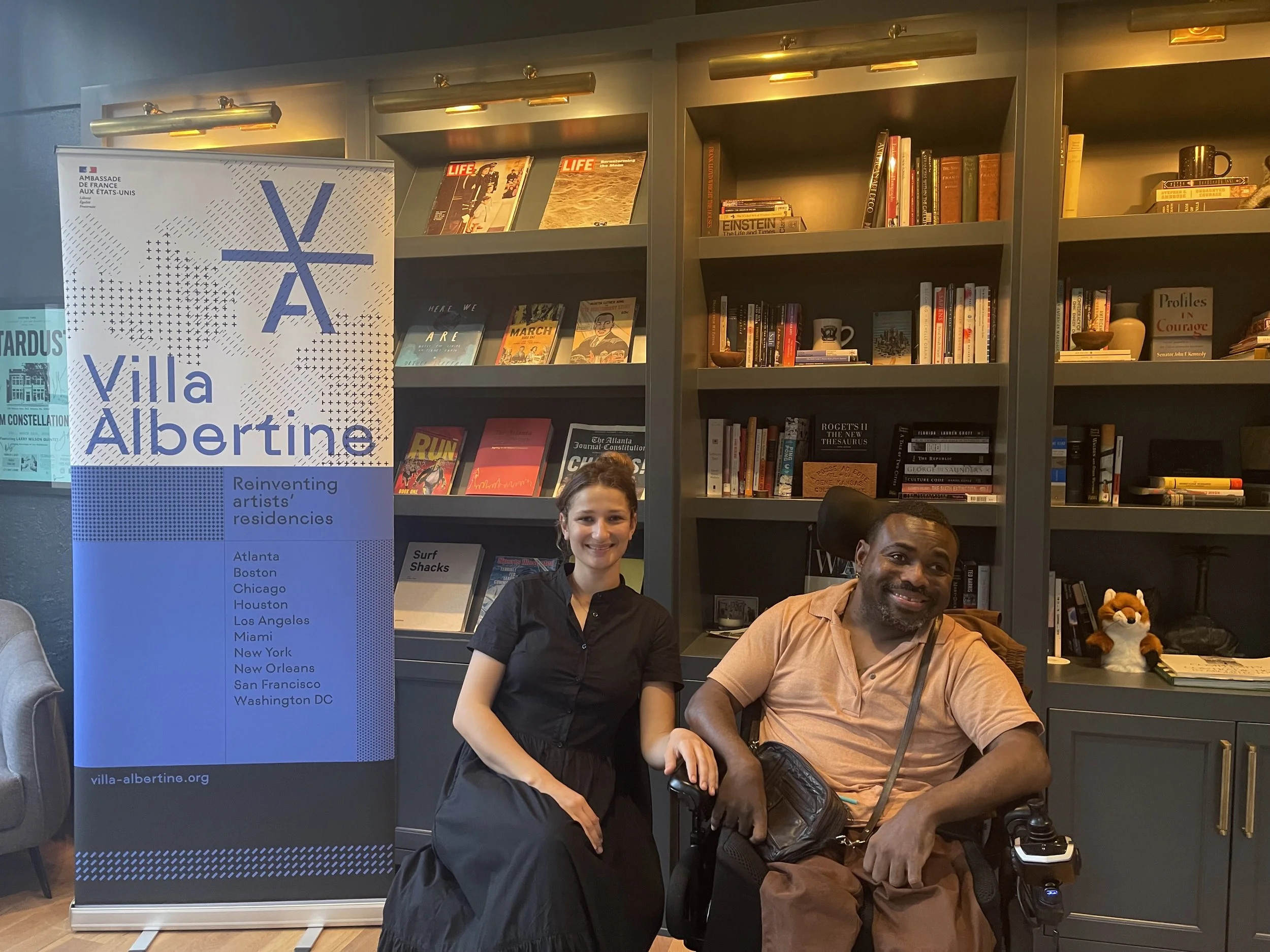 A smiling woman in a long black dress sits on a folding chair next to a man in a power wheelchair wearing a peach shirt. They are in a library with bookshelves behind them and a "Villa Albertine" banner listing American cities to the left.