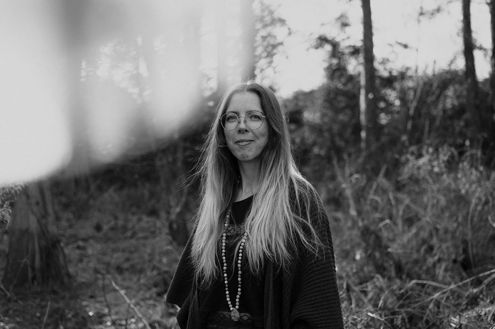A woman with long hair and glasses standing outdoors in a wooded area, looking at the camera.