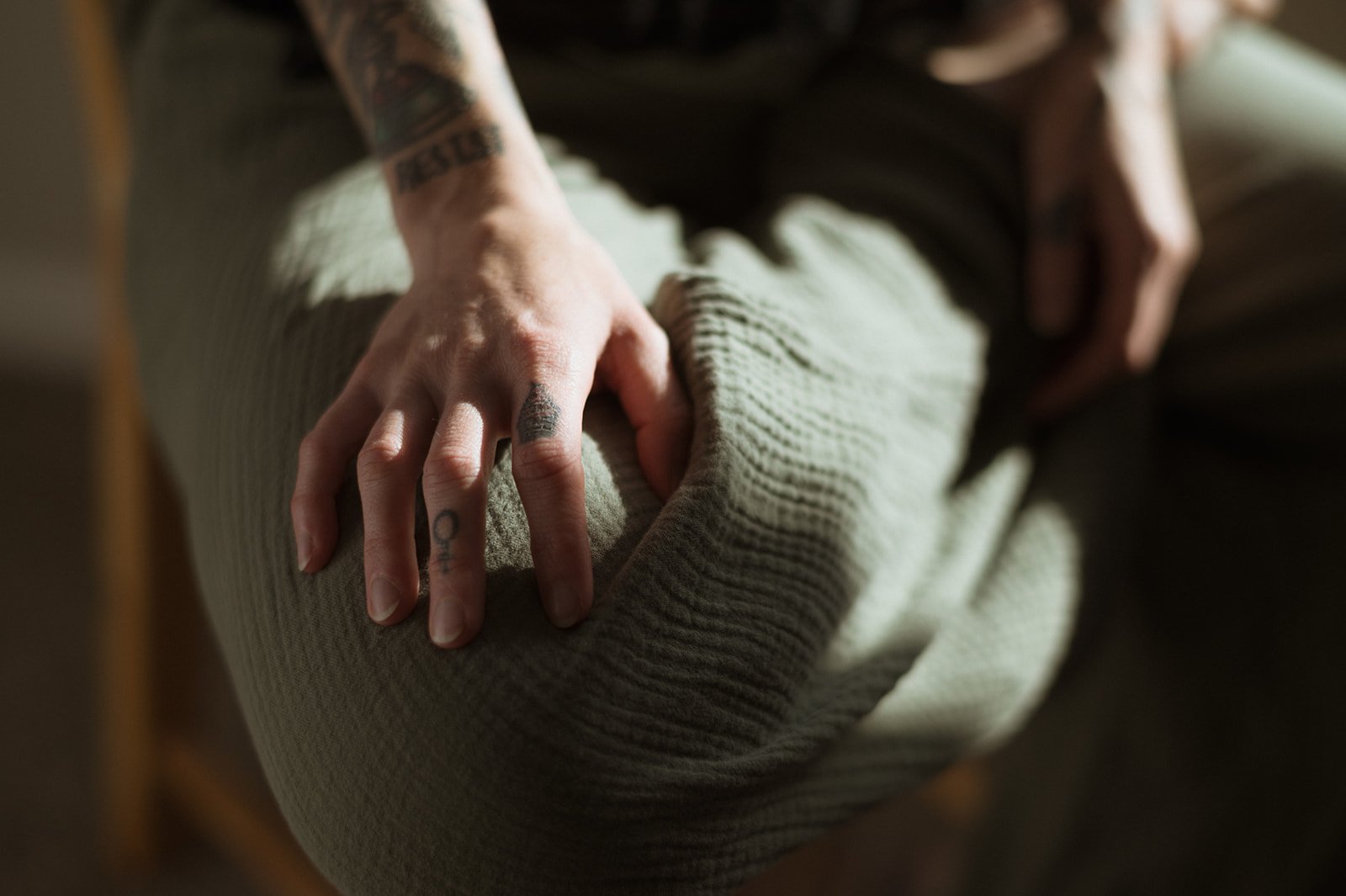 Close-up of a person's hand resting on their knee, with tattoos visible on fingers and wrist, wearing a beige ribbed knit sweater.