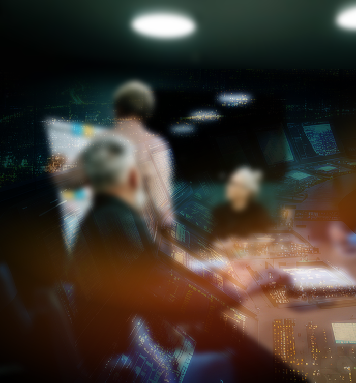 Blurred image of people working at computer monitors in a control room with multiple screens and a dark environment.