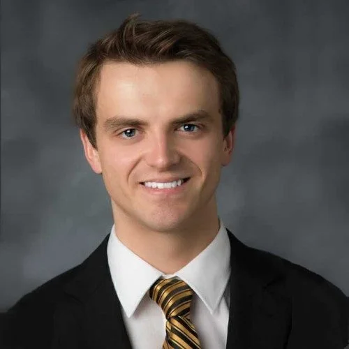 A young man in a suit and striped tie smiling against a gray background.